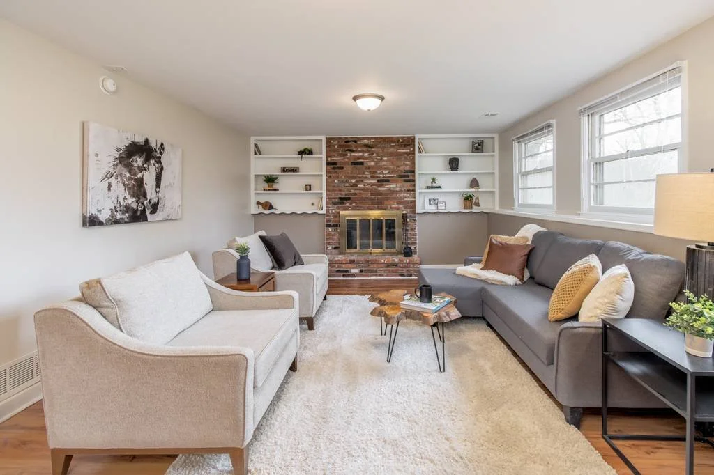 Living room with a beige sofa, gray sectional with throw pillows, white built-in shelves, brick fireplace, and windows.