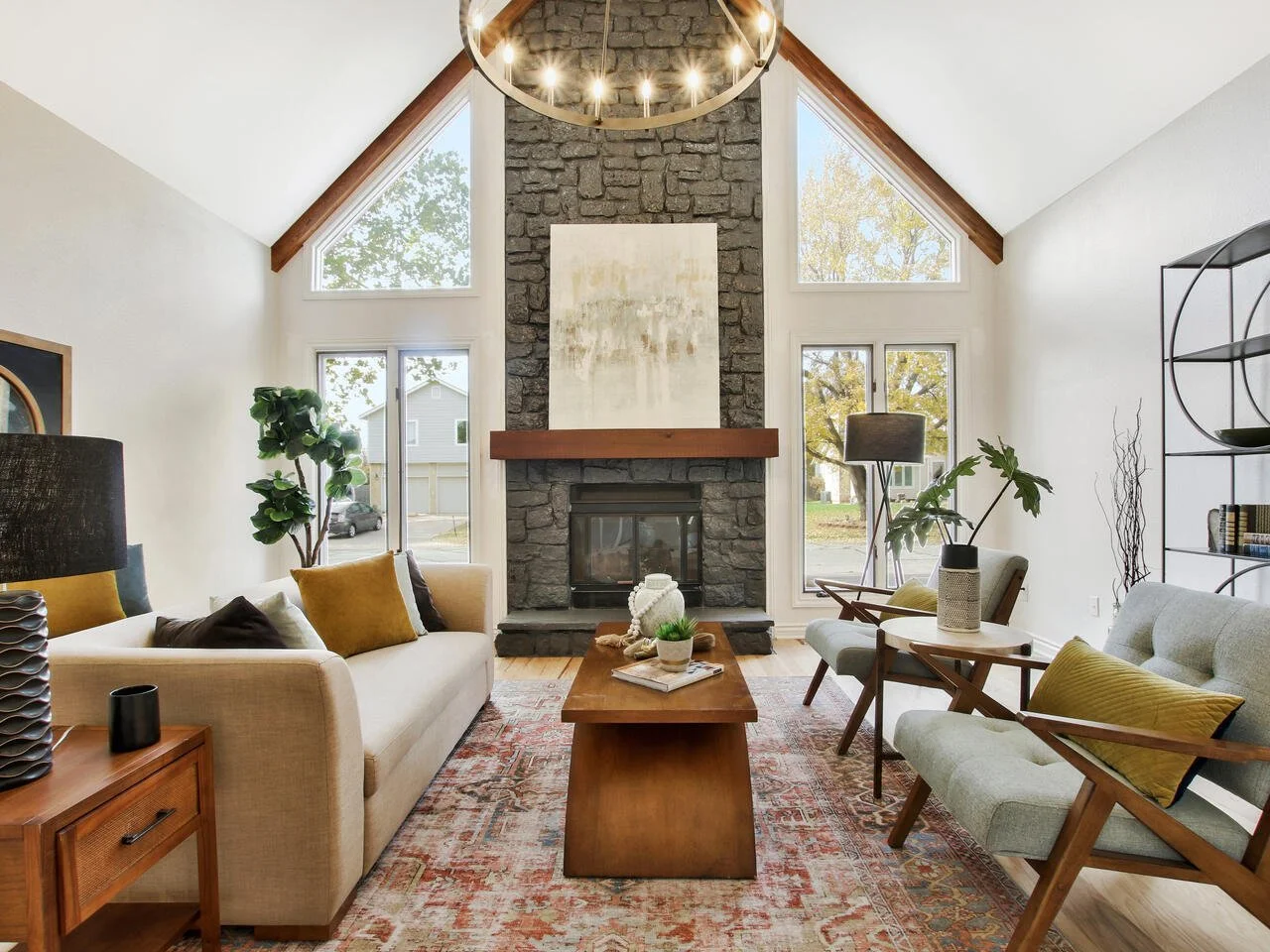 Living room with high vaulted ceiling, large windows, stone fireplace, modern chandelier, beige sofa, wooden coffee table, and various chairs with throw pillows, natural light, and interior decor.