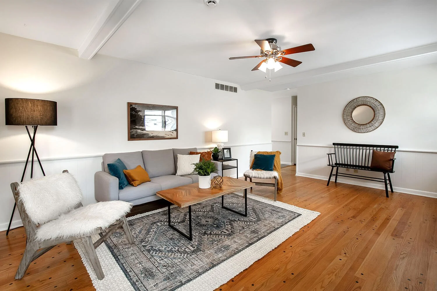 Living room with hardwood floor, white walls, and modern furniture including a gray sofa, a black bench with a brown pillow, a wooden chair with a white furry cushion, and a white furry armchair. There are decorative pillows, a wooden coffee table, a black side table with a lamp and picture frame, and a large black rug. The room has a ceiling fan with wooden blades, a large floor lamp, and wall art, including a round mirror and a framed picture.