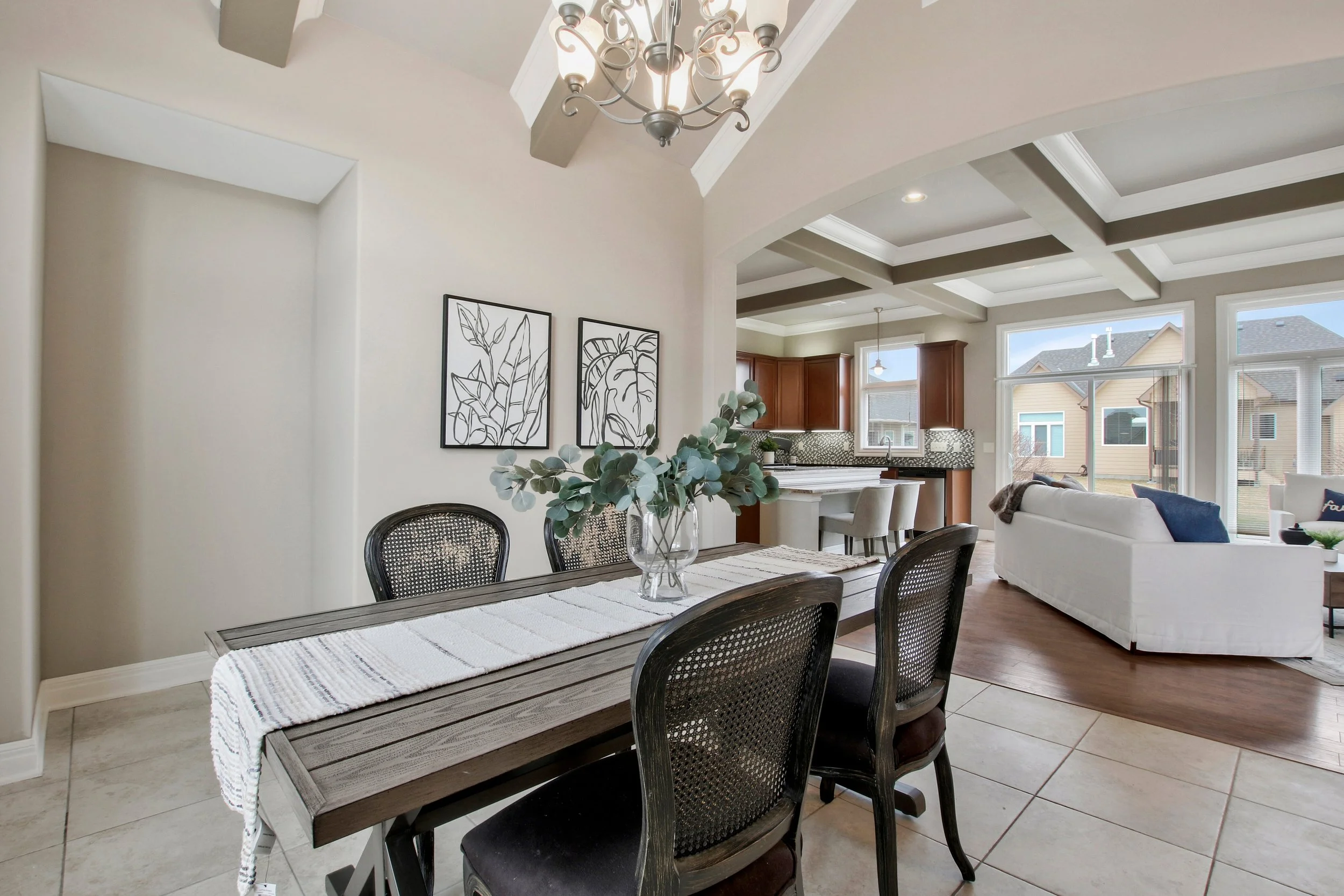 Open-concept dining and living room with large windows, light-colored walls, a wooden dining table with four chairs, and a bright kitchen with wooden cabinets in the background.