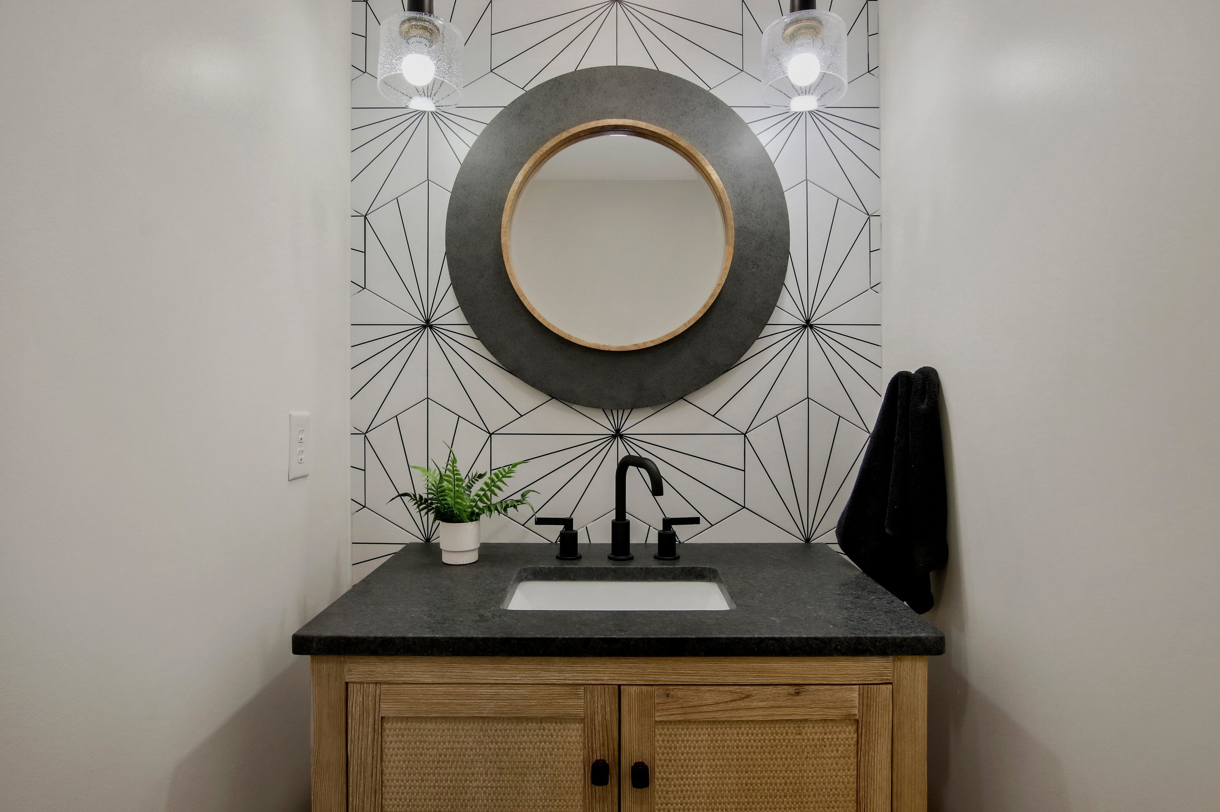 A bathroom vanity with a black countertop, a white sink, and a black faucet. There is a small potted green plant to the left of the sink. Above the mirror is a large circular mirror with a black and wood frame. The wall behind the mirror features geometric black lines on a white background. There are two pendant lights hanging above the mirror and a black towel hanging on the right side of the wall.