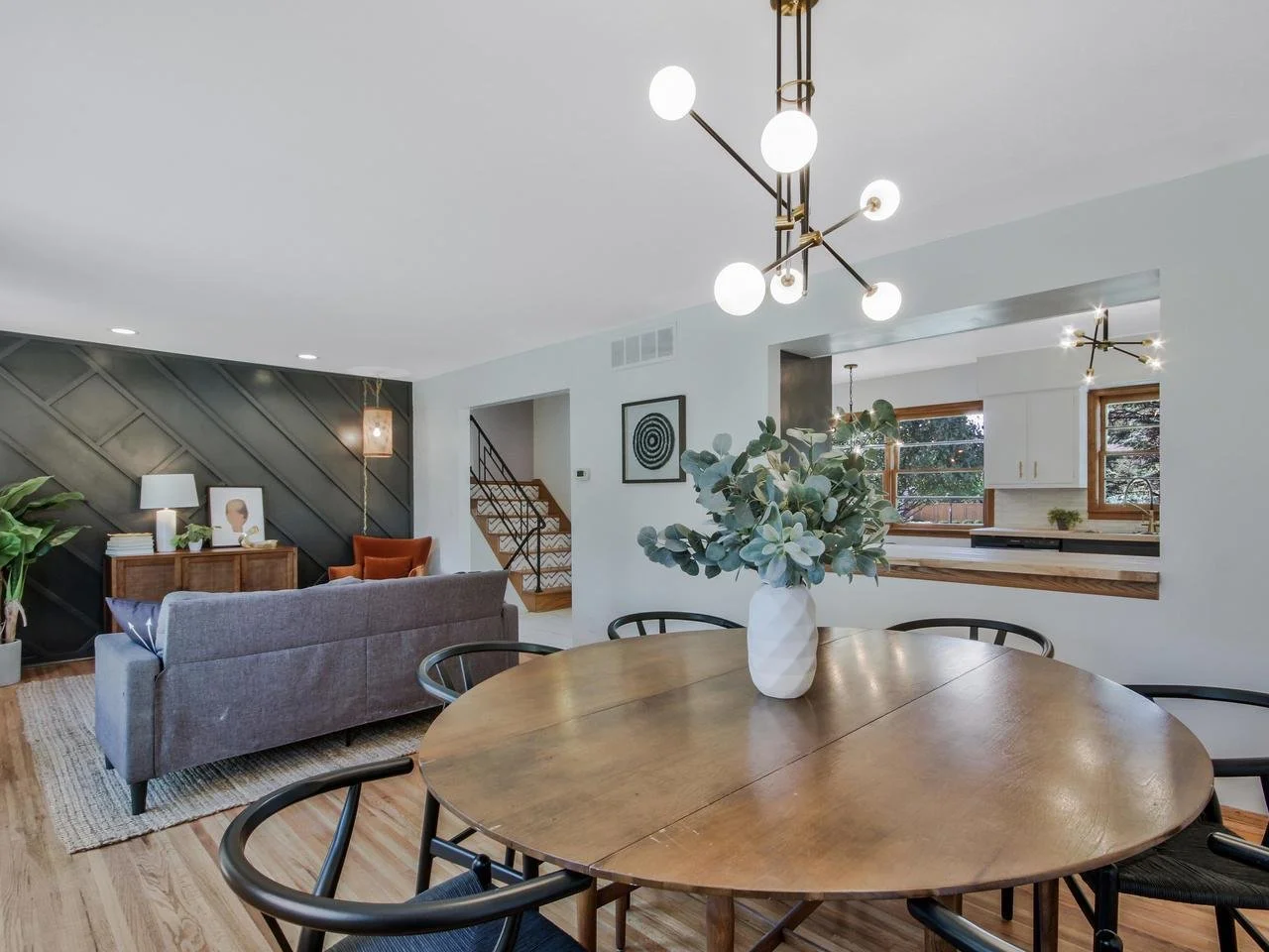 Open-concept living and dining area with modern design, wooden flooring, a round wooden dining table with a white vase of eucalyptus, gray and orange seating, and stylish lighting fixtures.