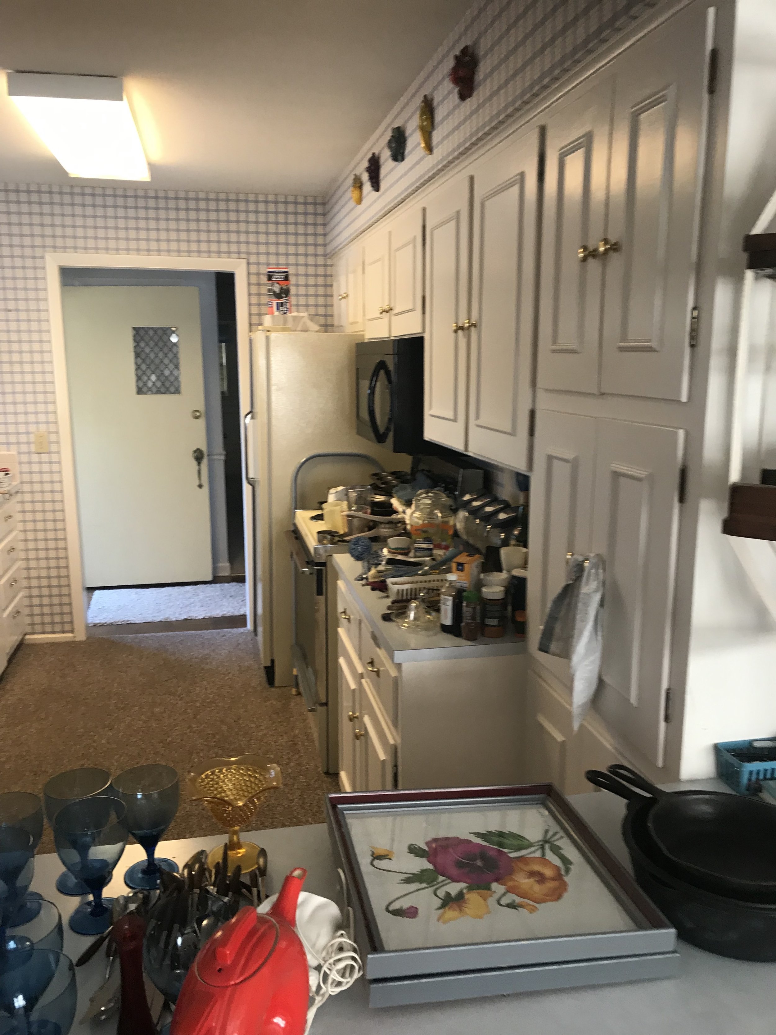 Cluttered kitchen counter with glasses, utensils, and a red iron. Background shows white cabinets, a microwave, and a refrigerator. The wall has patterned wallpaper and decorative items on top of cabinets.