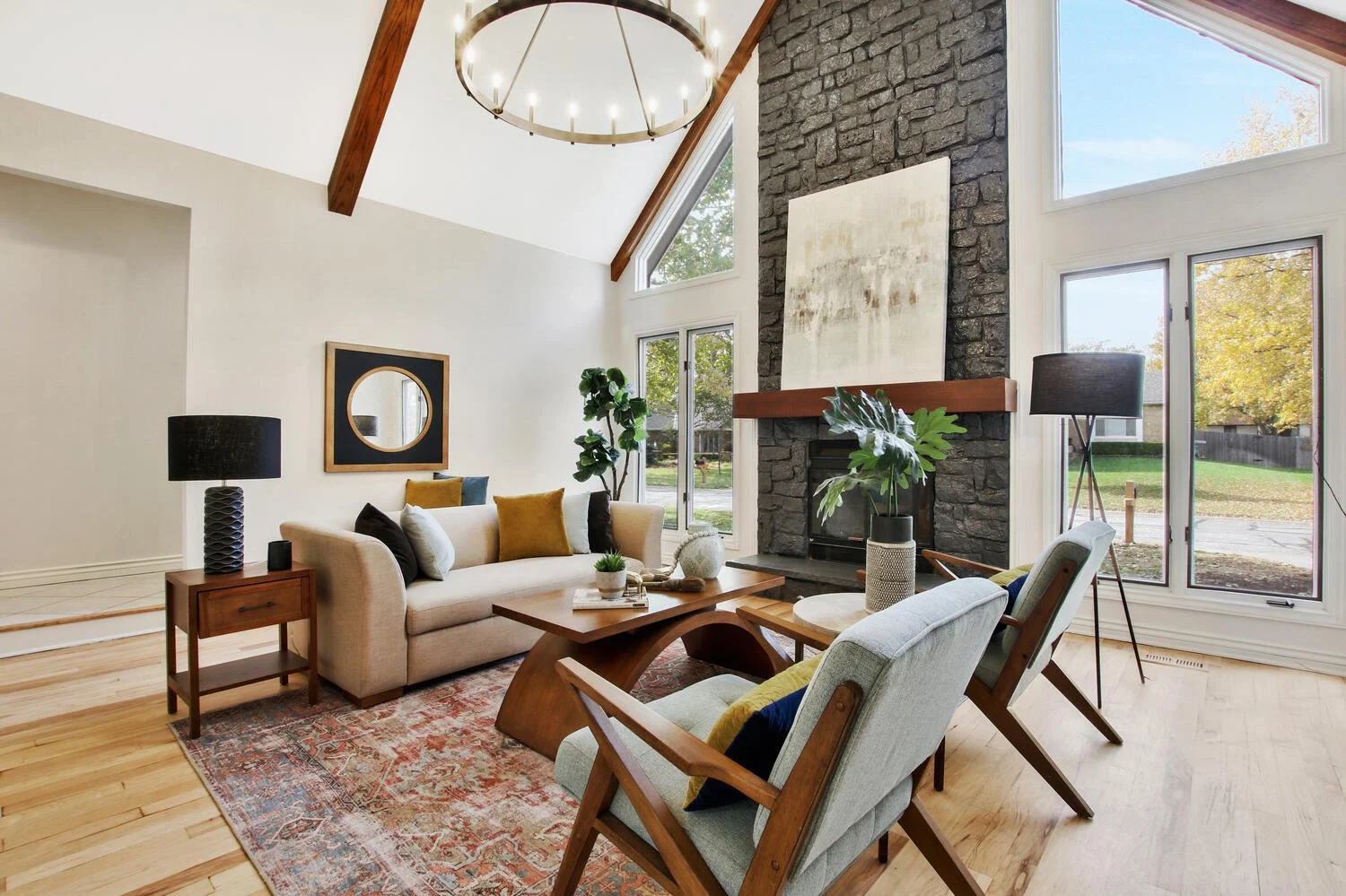 Living room with high vaulted ceiling, large windows, and a black stone fireplace. Furnished with a beige sofa, mid-century chairs, a wooden coffee table, and decor including pillows, plants, a lamp, and artwork.