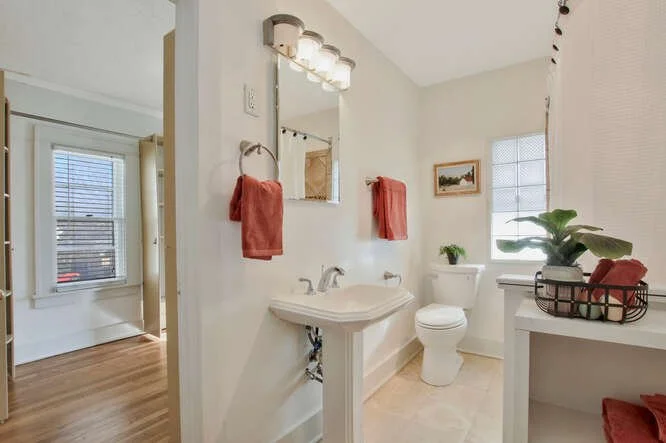 Bathroom with a pedestal sink, a toilet, a mirror above the sink, a window with blinds, and decorative red towels. There is a plant on the windowsill and a basket with a red cloth on a nearby table.
