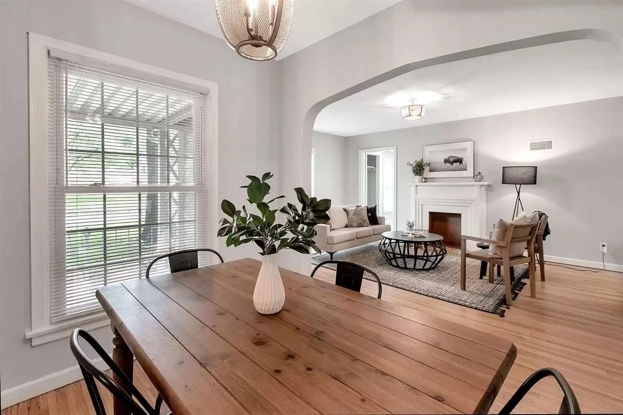 Open concept living and dining room with wooden floors, white walls, large window with blinds, and a central archway. The dining table has a vase with green leaves. The living room features a white sofa, a round coffee table, a fireplace with artwork above, and a mix of chairs and a floor lamp.