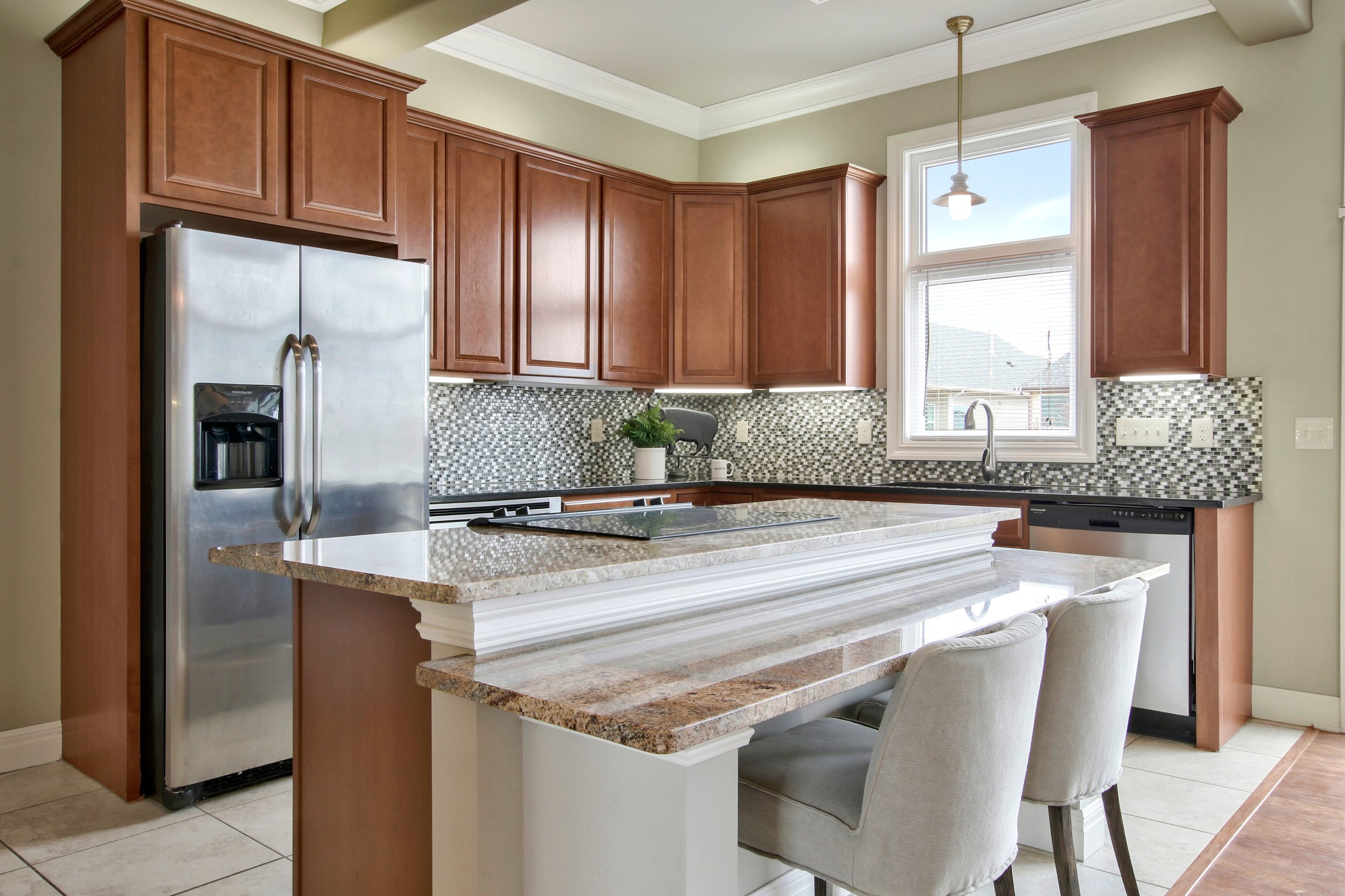 Kitchen with wooden cabinets, granite countertops, mosaic tile backsplash, stainless steel refrigerator, dishwasher, and a kitchen island with seating. A window above the sink lets in natural light.