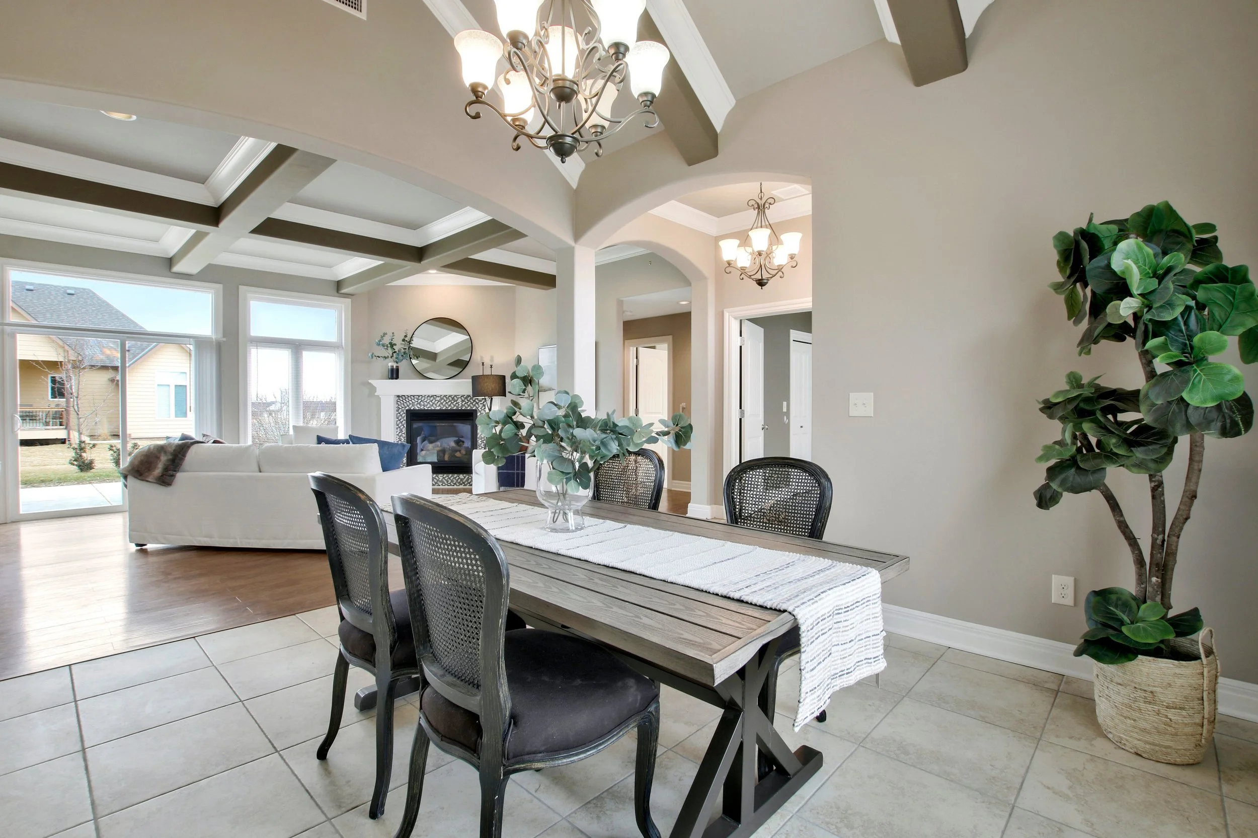 Open-concept living and dining area with a tiled floor, wooden dining table with chairs, a white sofa, a fireplace, and large windows bringing in natural light, decorated with chandeliers and potted plants.