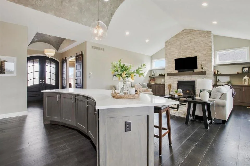 A modern, open-concept living room and kitchen area with dark wood floors, a curved kitchen island, a white fireplace with a mounted flat-screen TV, beige sofas, and decorative shelves with various items.