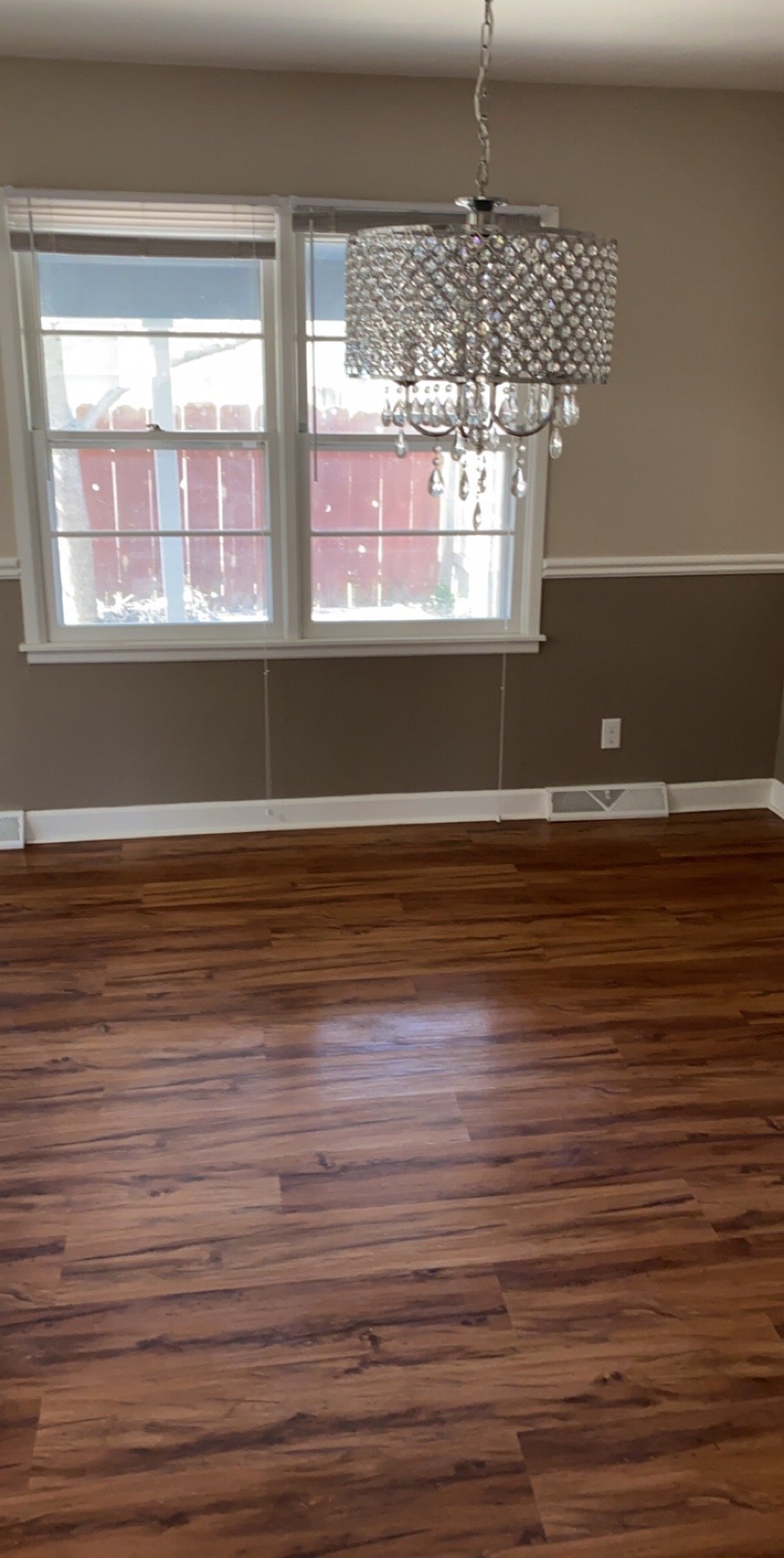 Empty room with wooden floor, beige walls, a window overlooking a backyard, and a crystal chandelier hanging from the ceiling.
