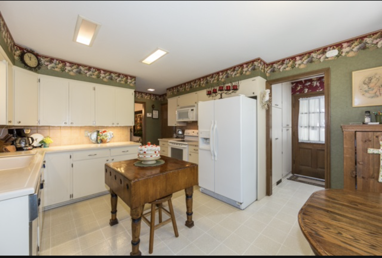 A cozy kitchen with white cabinets, a wooden table with a cake, green and floral wallpaper, and a wooden door with a window.