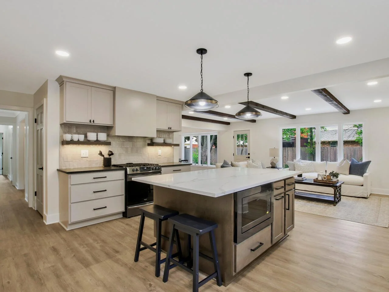 Open-concept kitchen and living room with white walls, wood flooring, and large windows. The kitchen features a central island with a microwave, pendant lights, and a stove. The living area has a white sofa with cushions, a coffee table, and a rug.