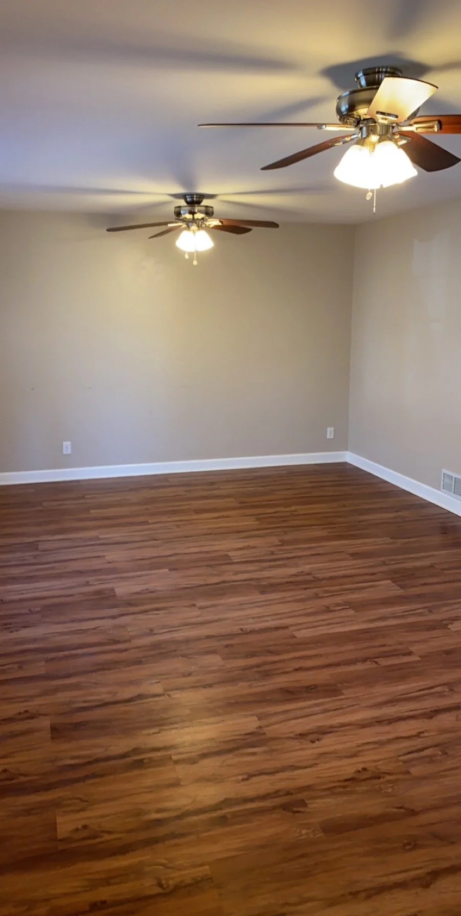 Empty room with hardwood floors, beige walls, white baseboards, two ceiling fans with lights, and a vent on the lower right wall.