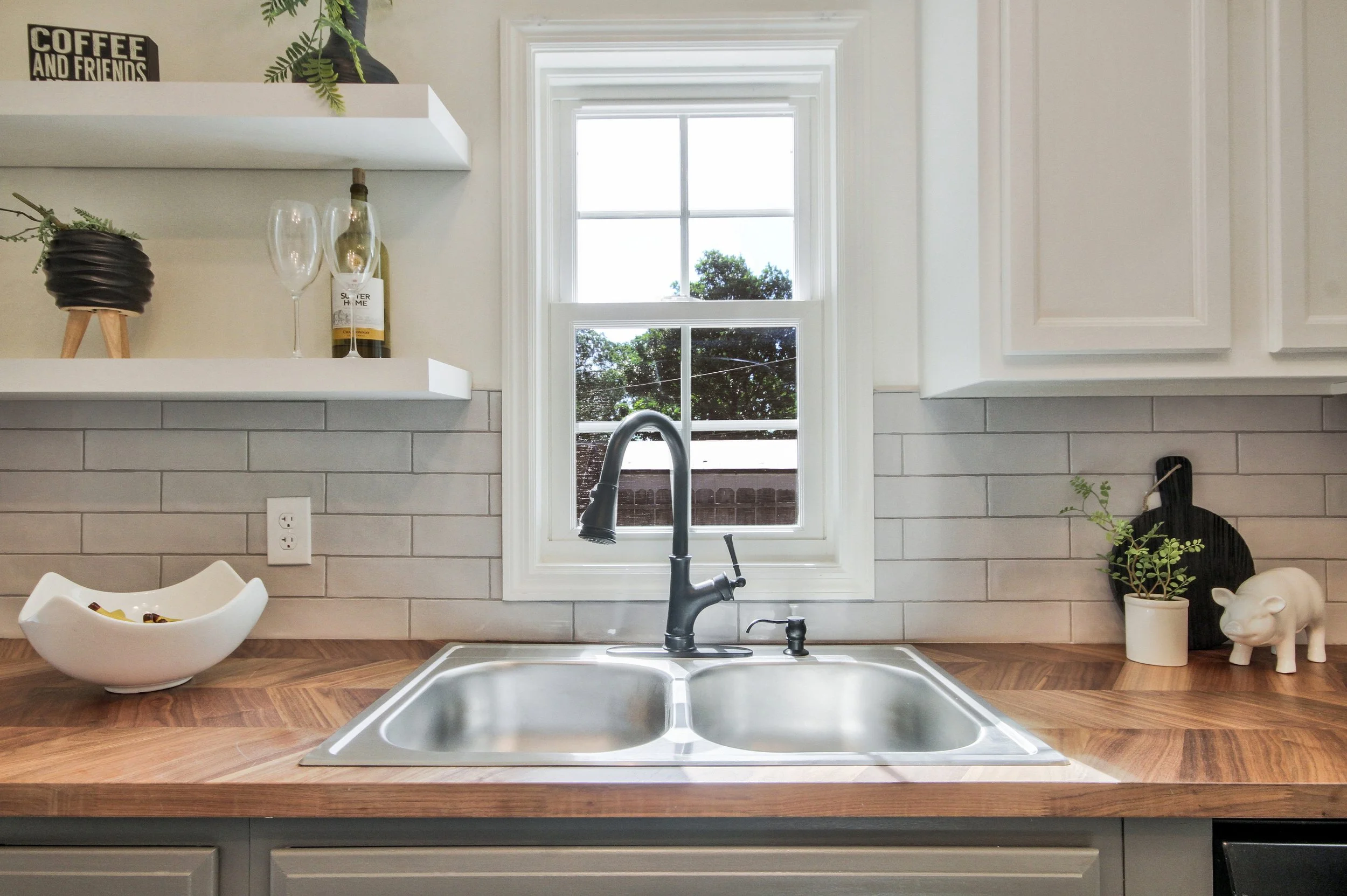 Kitchen sink with a black faucet, window with greenery outside, white cabinets, gray tiled backsplash, wooden countertop with a decorative bowl, plant, black cutting board, and ceramic pig figurine.