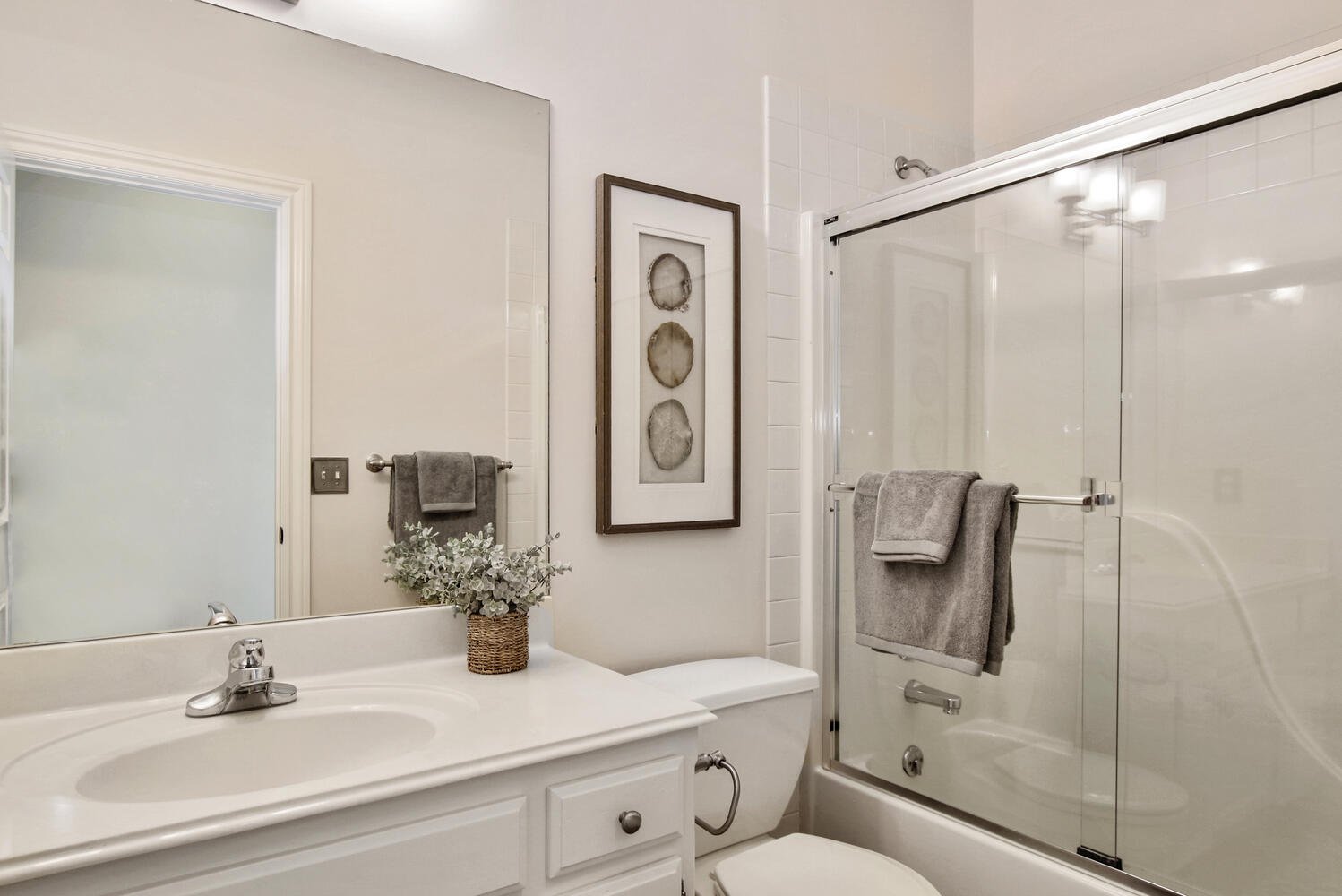 A clean bathroom with a white vanity sink, a large mirror, a framed artwork with three circular stone designs, a potted plant, a glass shower with a sliding door, and gray towels hanging on towel bars.