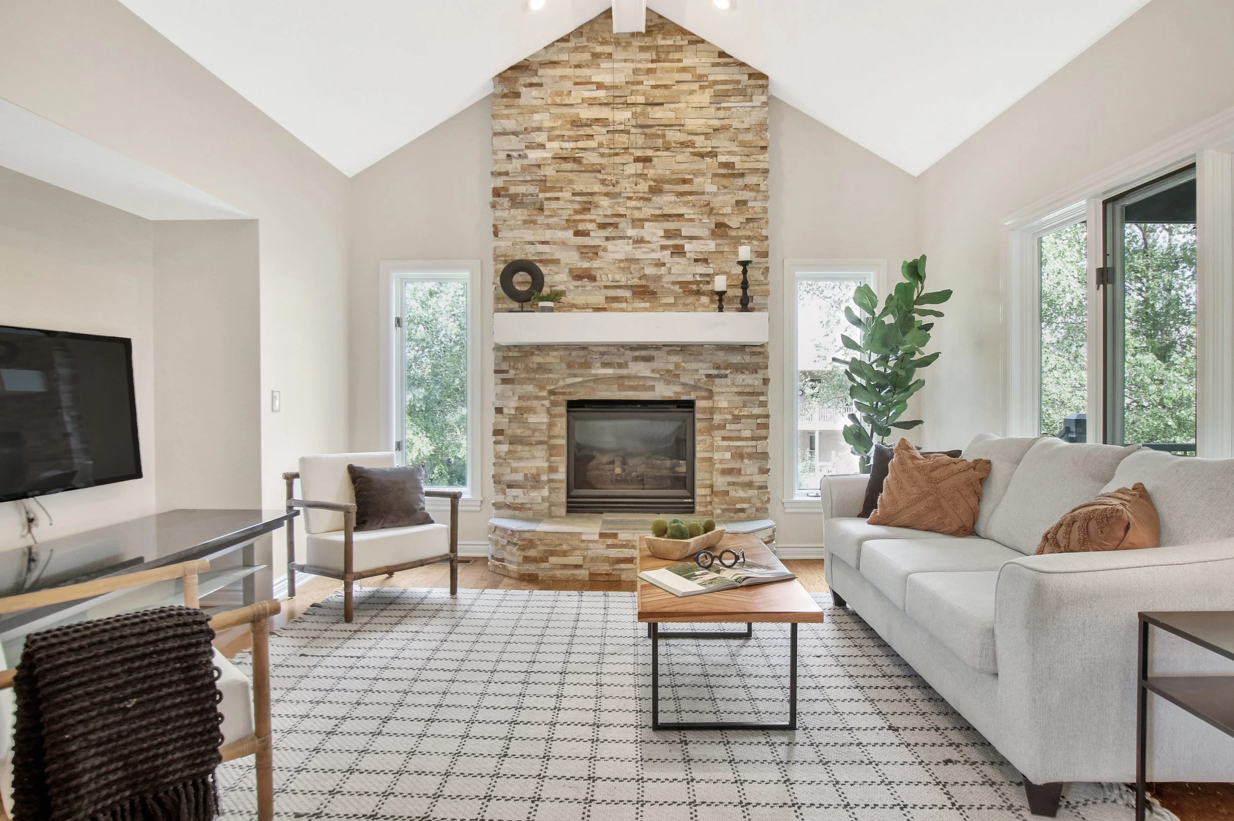 Living room with a fireplace, white couch with pillows, wooden coffee table, armchair, large windows with green trees outside, and a patterned rug.
