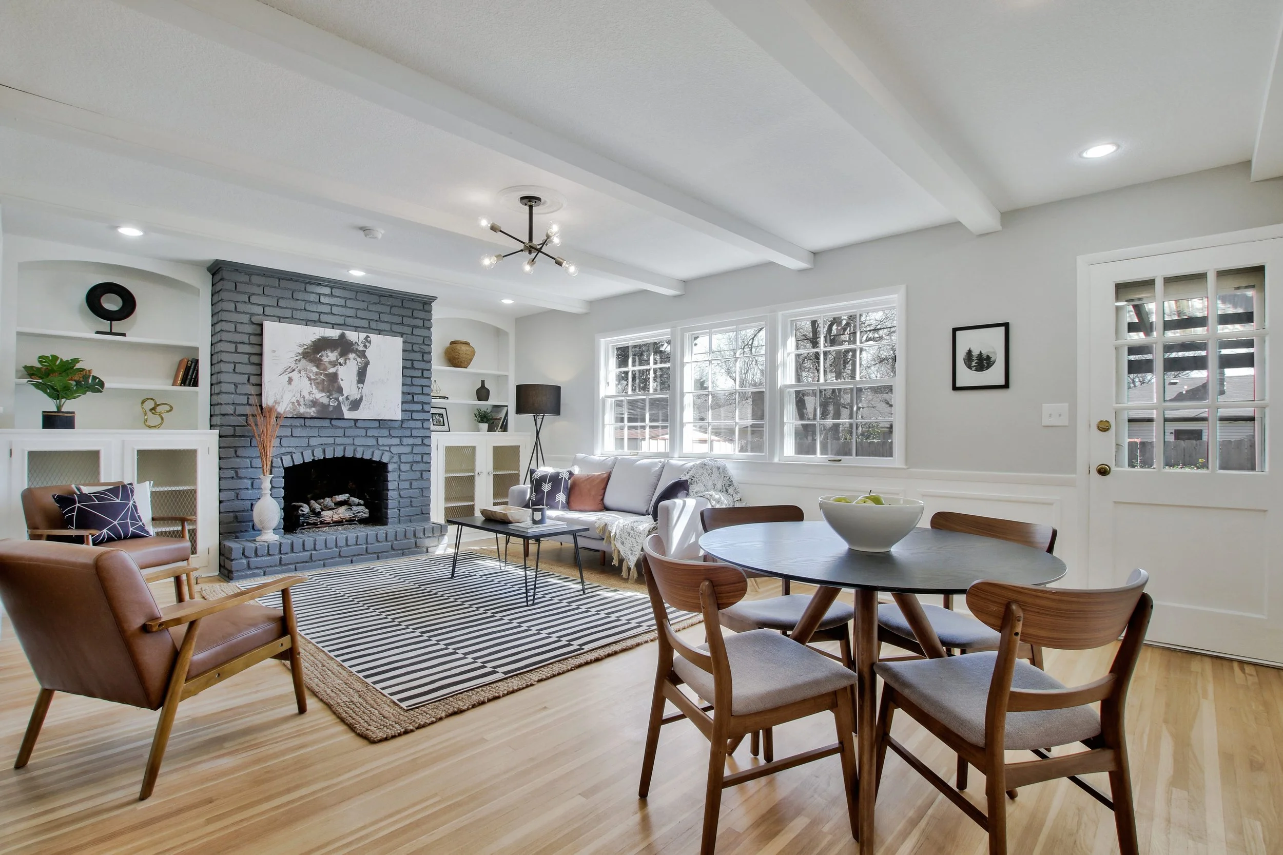 Bright living room with sleeping area, featuring a black brick fireplace, white cabinetry, and a round dining table with four wooden chairs. Large windows let in natural light, and decor includes plants, art, and a patterned rug.