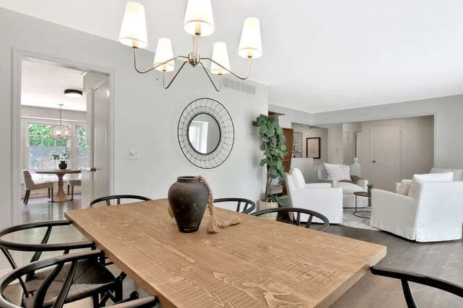 Dining room with a wooden table, black chairs, a modern chandelier, and a mirror on the wall; living room with white sofas and a door leading to an outdoor patio.