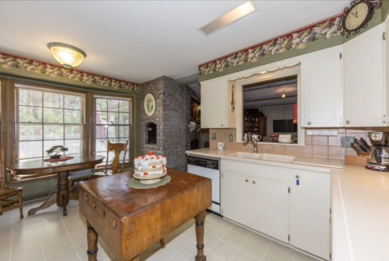 Kitchen with white cabinets, a wooden table with a cake, a window with blinds, and a fireplace with a stone chimney.