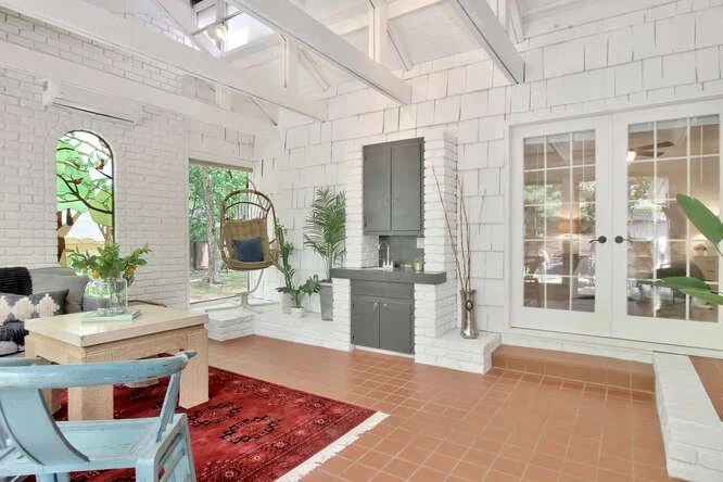 Bright conservatory with white painted brick walls, large windows, and terracotta tiled floor. Contains a hanging chair, houseplants, a side table, and a red area rug.