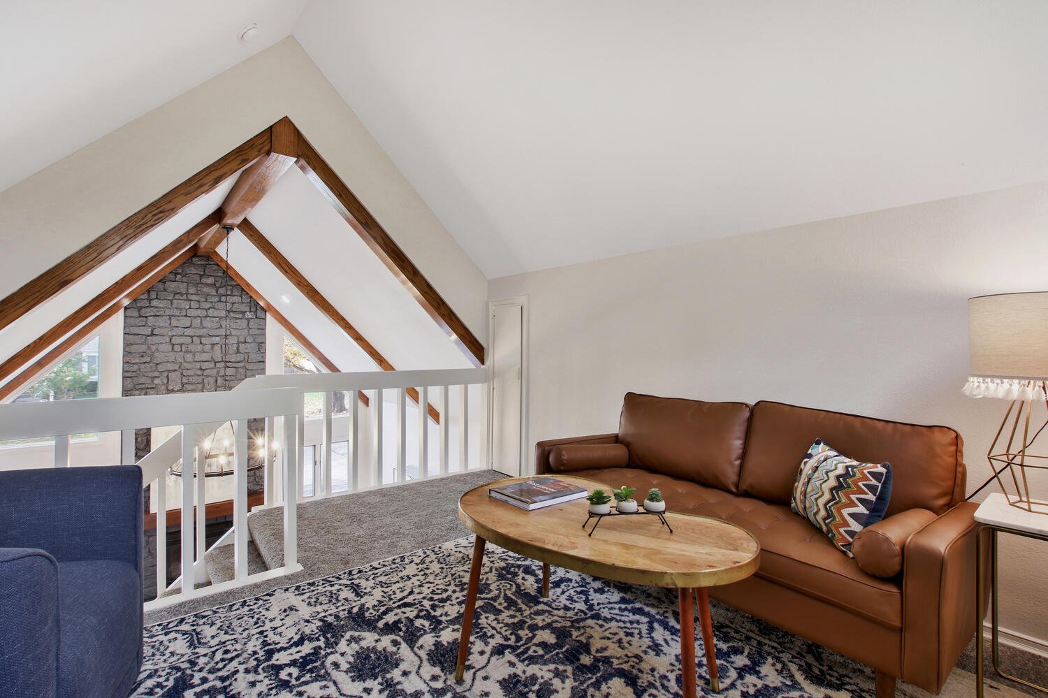 Living room with a brown leather sofa, patterned cushion, wooden coffee table with books and small plants, white side table with lamp, patterned rug, and angled ceiling with wood beams and large windows with brick wall outside.