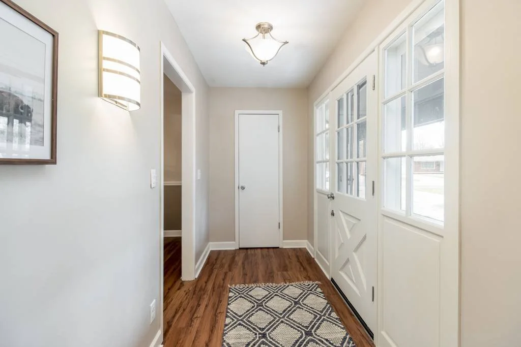 Entryway with cream-colored walls, a wooden floor, a patterned tile rug, a wall-mounted light, a framed picture, a white door, and a white door with windows leading outside.