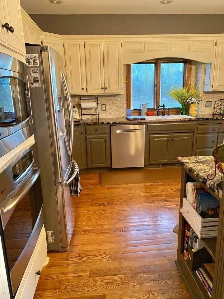 A kitchen with cream upper cabinets and green lower cabinets, a window above the sink, and hardwood floors.