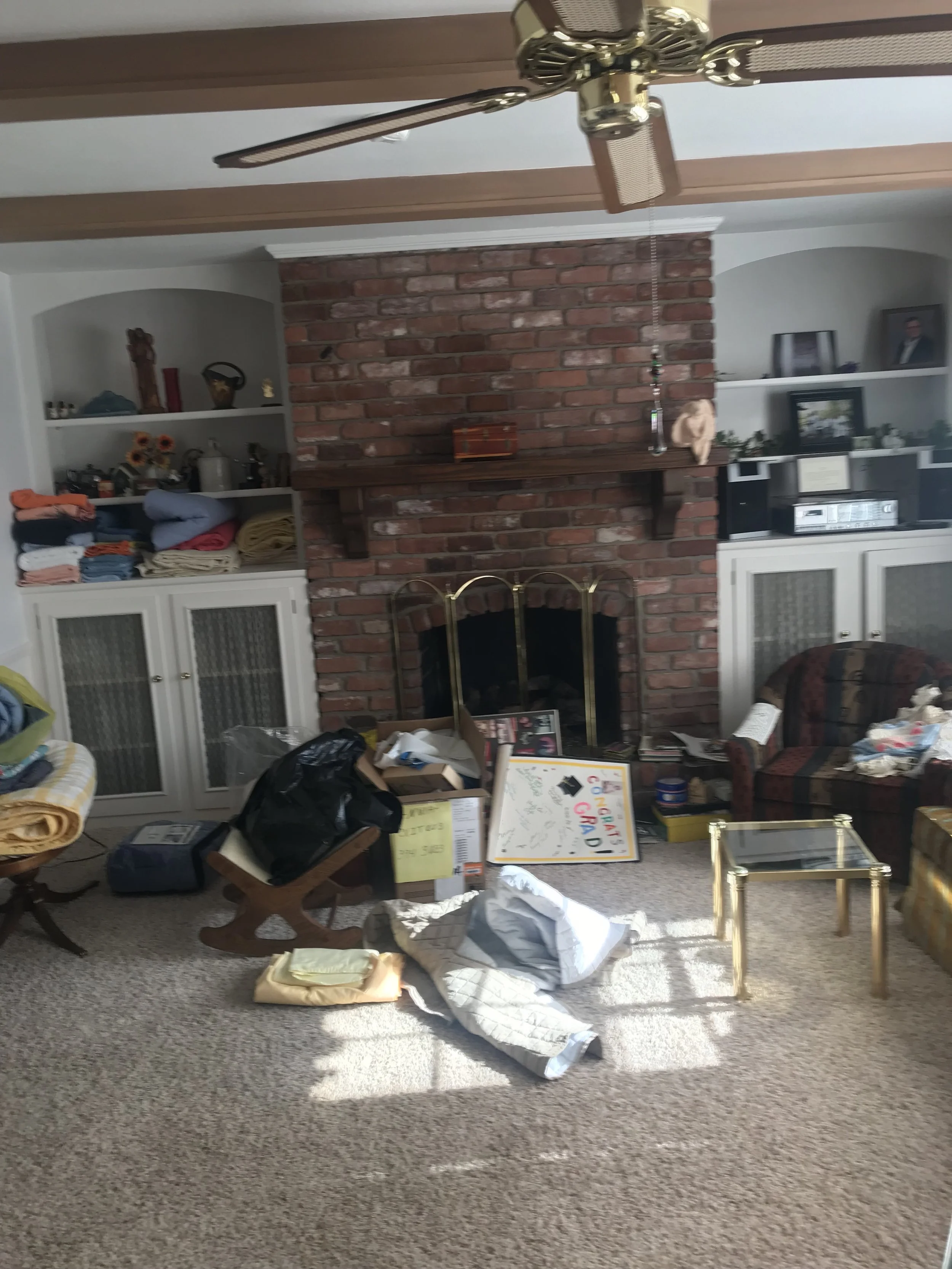 Living room with brick fireplace, white built-in shelves on both sides, a ceiling fan, and various objects and clothes scattered around.