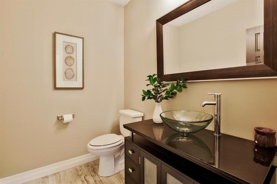 Bathroom with beige walls, a white toilet, a dark vanity with a glass vessel sink, a round mirror, a potted plant, and wall art.