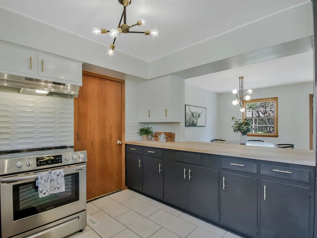Modern kitchen with white upper cabinets, gray lower cabinets, wooden countertop, stainless steel stove, wood door, artwork on the wall, and pendant lighting fixtures.