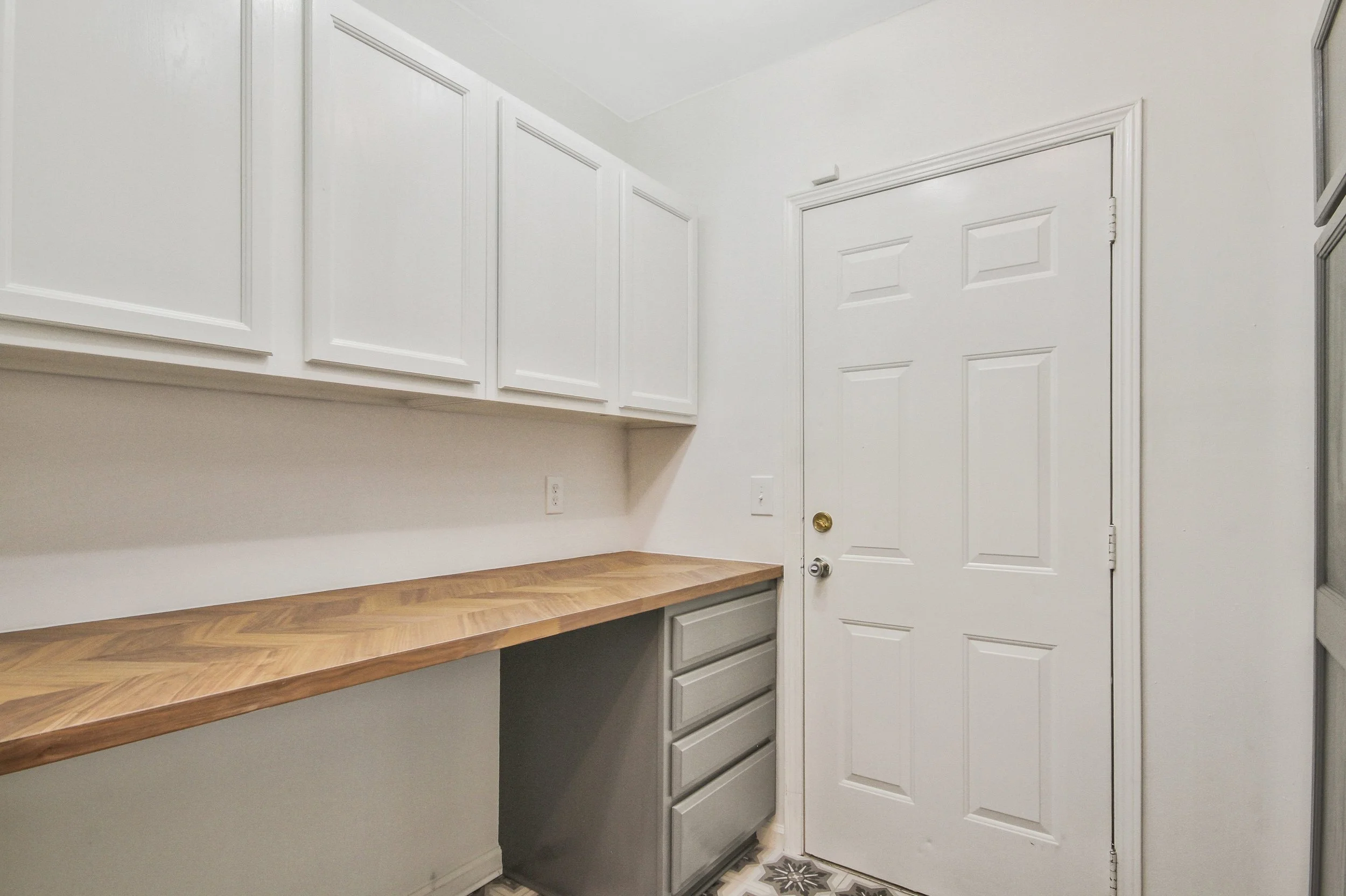 Small laundry room with white cabinets, a wooden countertop, and a white door, with a patterned tile floor.