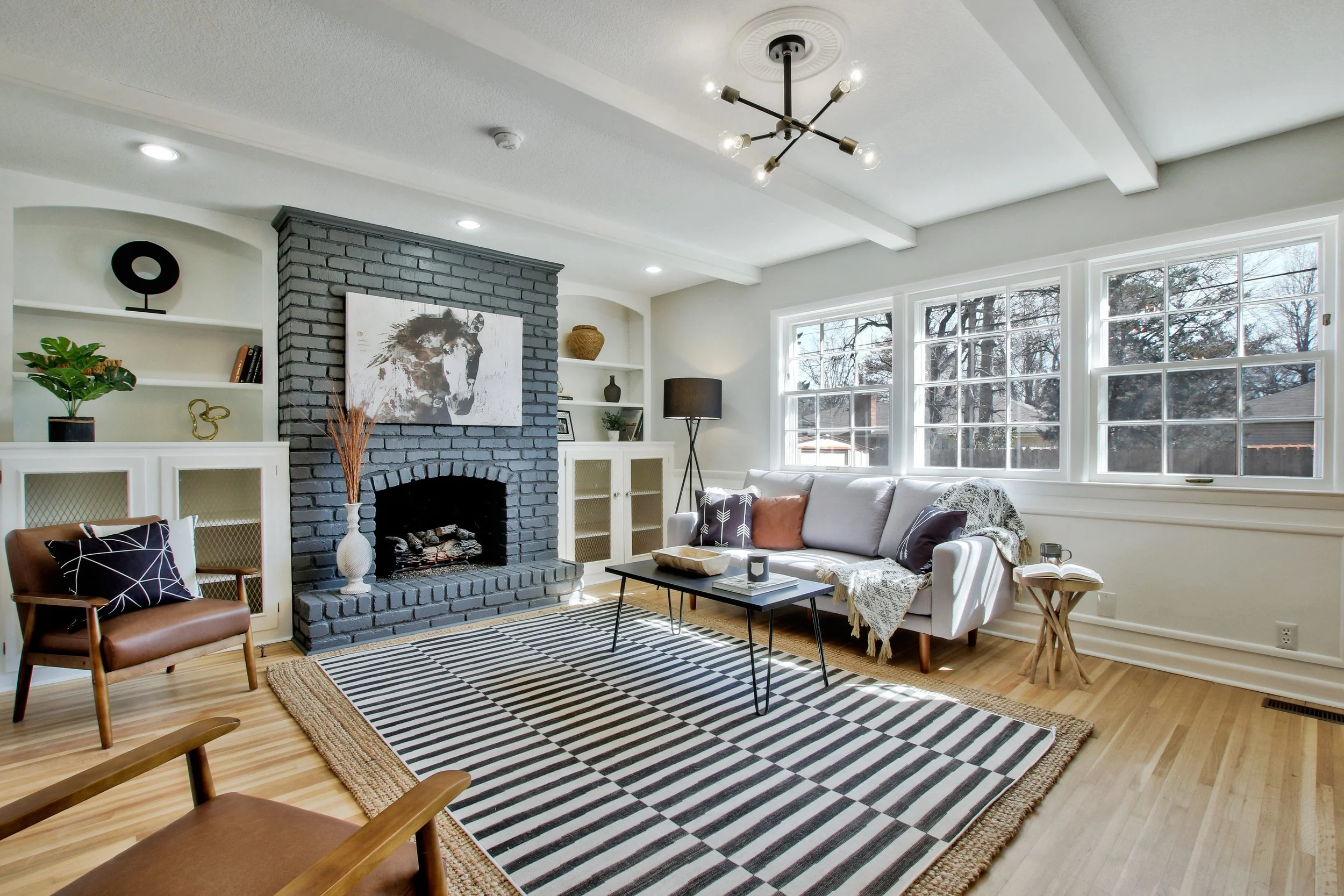 Bright living room with large windows, white walls, a black brick fireplace, a geometric black-and-white rug, a white sofa with accent pillows, a wood armchair with a pillow, and modern lighting fixtures.