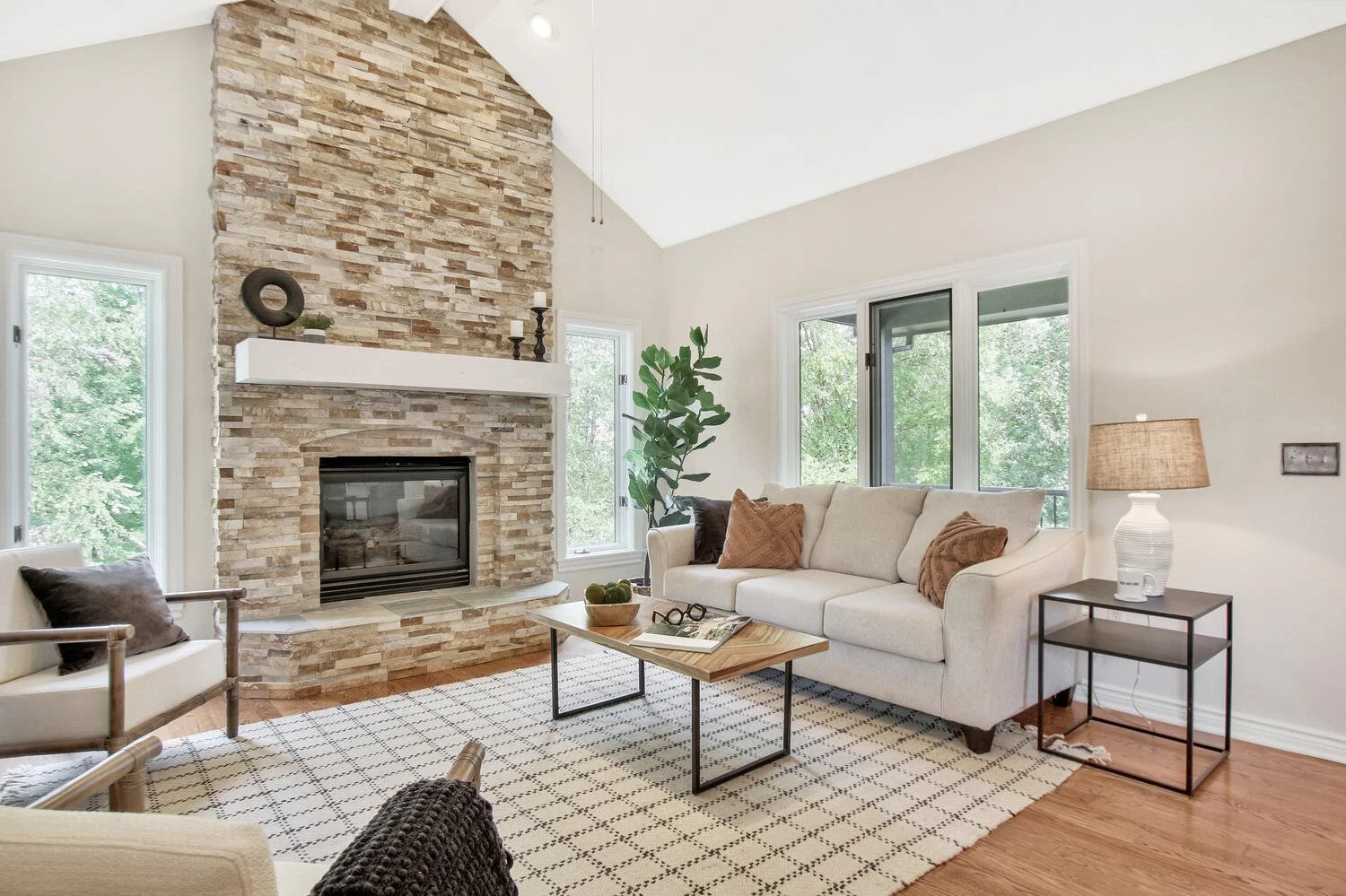 Living room with beige sofa, wooden coffee table, stone fireplace, large windows, and lamp on side table.