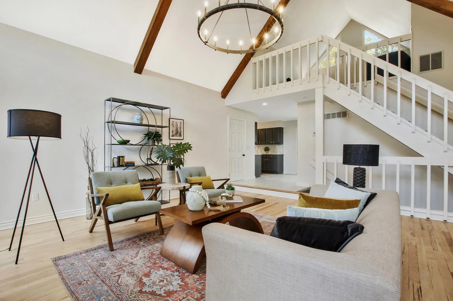 Living room with beige sofa and two armchairs, wooden coffee table, black floor lamp, black bookshelf, and a white staircase with wooden railing leading to an upper floor. The room has vaulted ceiling with exposed wooden beams, chandelier, and light-colored hardwood flooring.