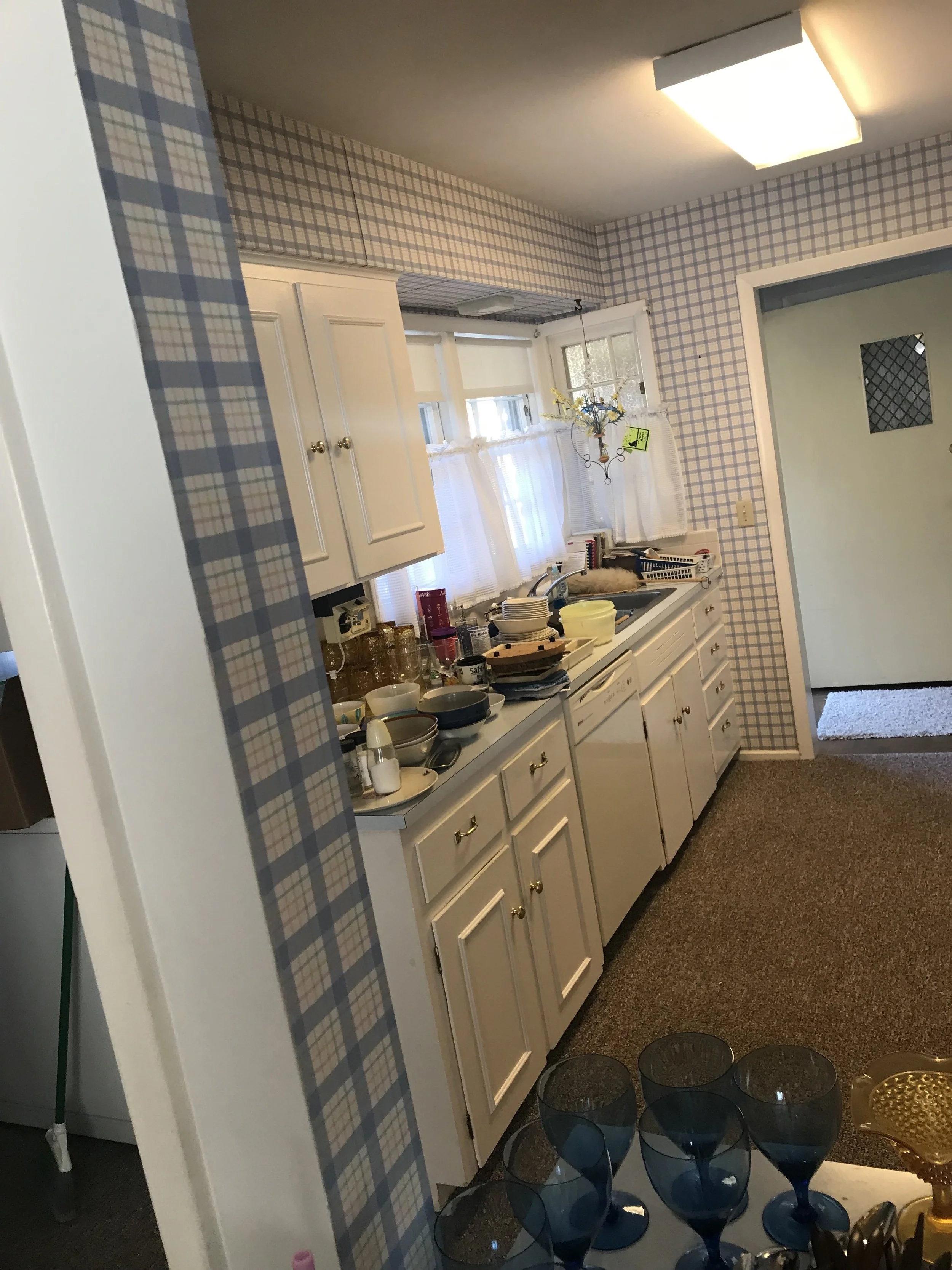 A kitchen with white cabinets, a window with white curtains, and a cluttered countertop. There is a light fixture on the ceiling and a door leading outside. Blue plaid wallpaper borders the room.