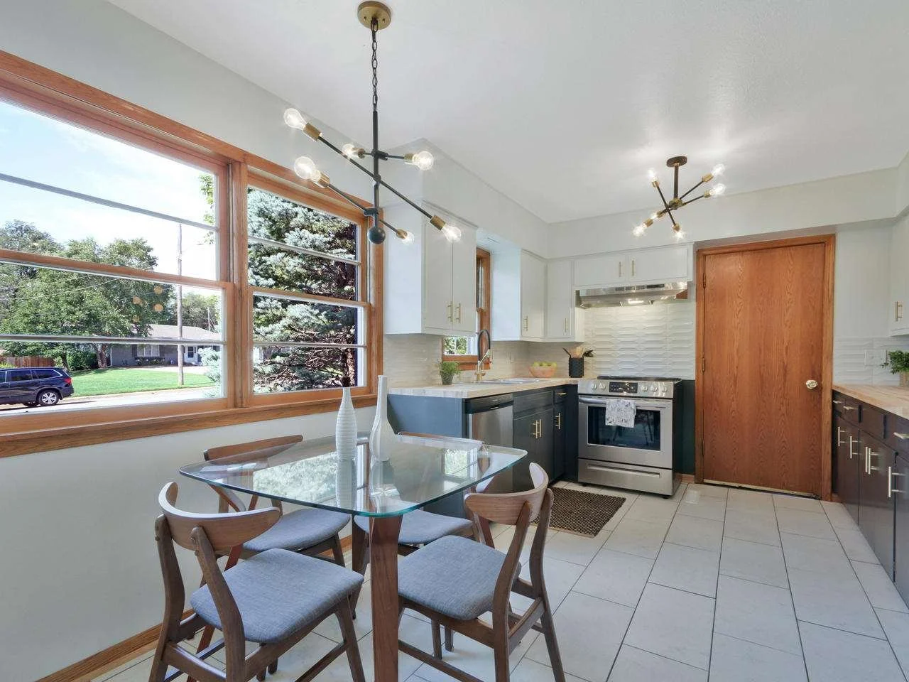 A bright kitchen with large windows, white and dark blue cabinets, stainless steel appliances, a glass dining table with four chairs, and modern lighting fixtures.