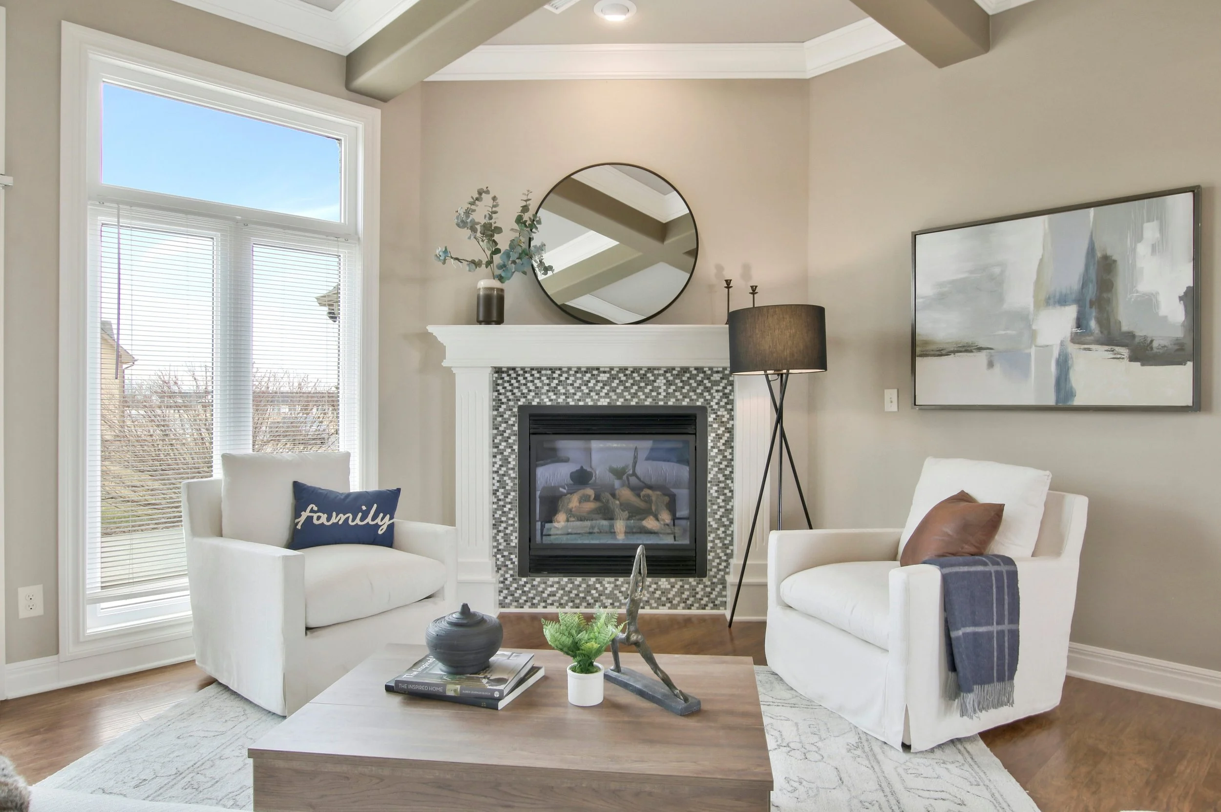 Living room with white armchairs, a fireplace with a round mirror above, a wooden coffee table with decor, large windows with blinds, and modern artwork on the wall.