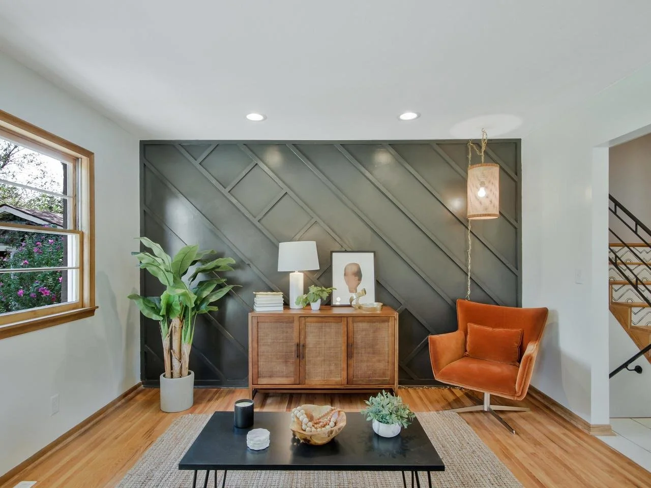 Living room with a large window, a wooden sideboard, a tall potted plant, a black coffee table, an orange armchair, a white lamp, and decorative items.