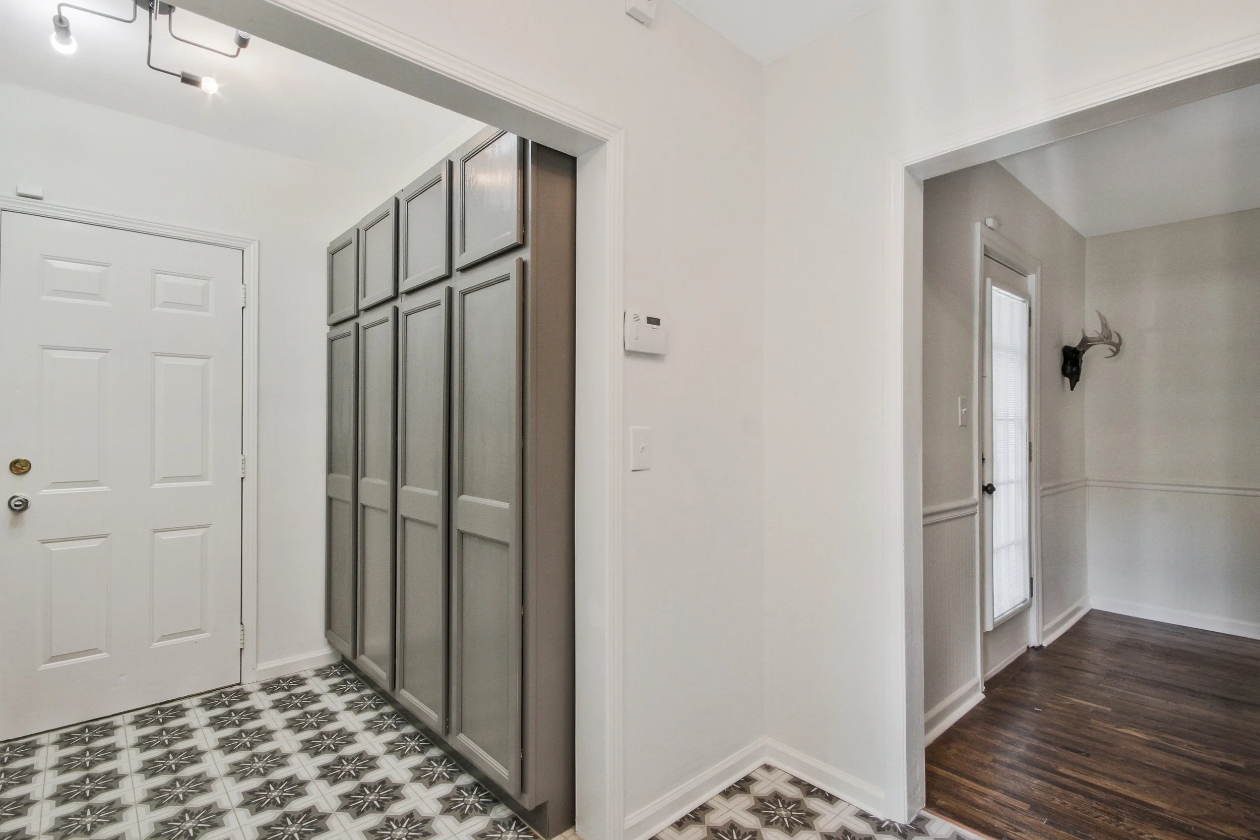 Entryway with white door, gray storage cabinets, patterned tile floor, and connecting hallway with wooden floor and a mounted deer head.