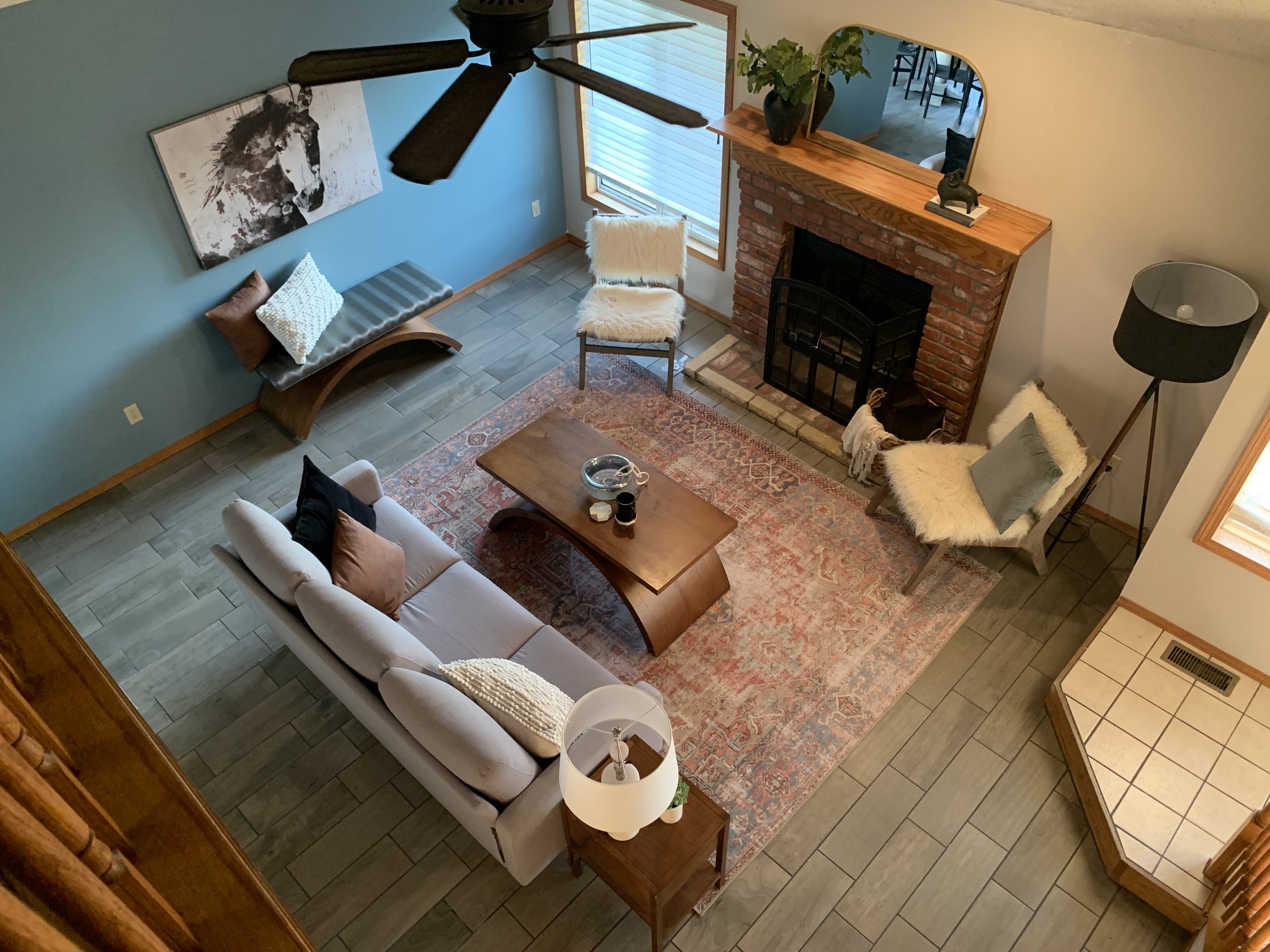 Living room with gray sofa, wooden coffee table, two cozy chairs with throw pillows and furry throws, brick fireplace with mirror above, large window with blinds, blue accent wall with horse painting, small side table with lamp, black floor lamp, and decorative items.