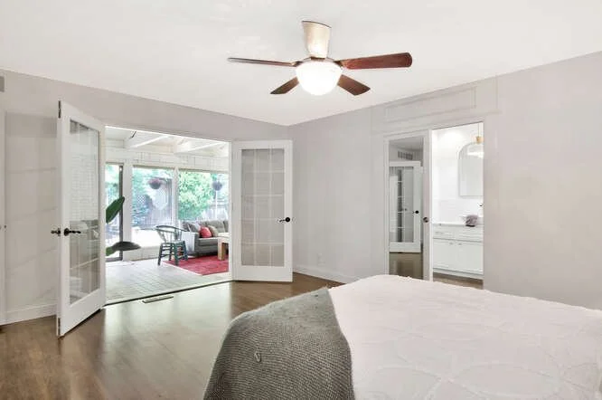 Empty bedroom with a ceiling fan, white walls, and hardwood floors, with doors leading to a bright sunroom with outdoor furniture.