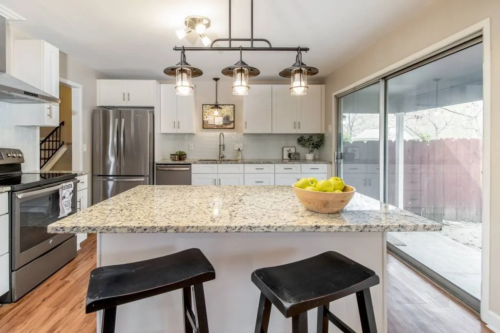 Modern kitchen with white cabinets, stainless steel refrigerator, granite island, black stools, and sliding glass door leading outside.