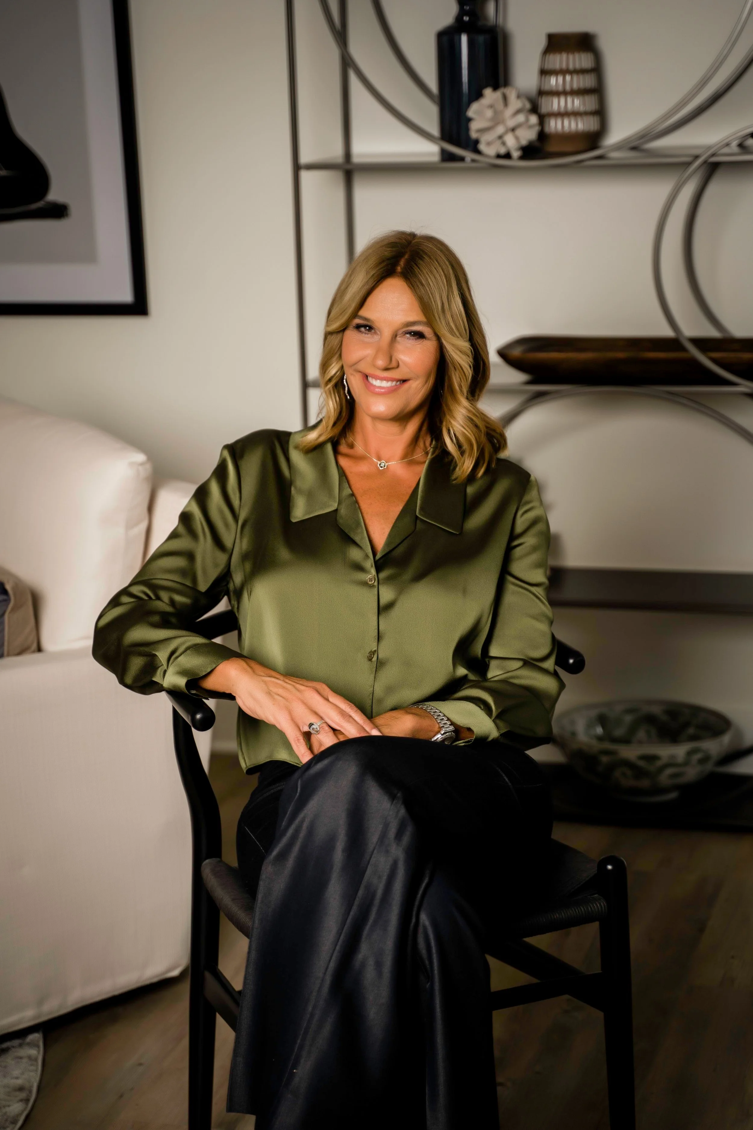 A woman with shoulder-length blonde hair, wearing a shiny green satin blouse and black pants, sitting on a black chair in a contemporary living room, smiling at the camera.