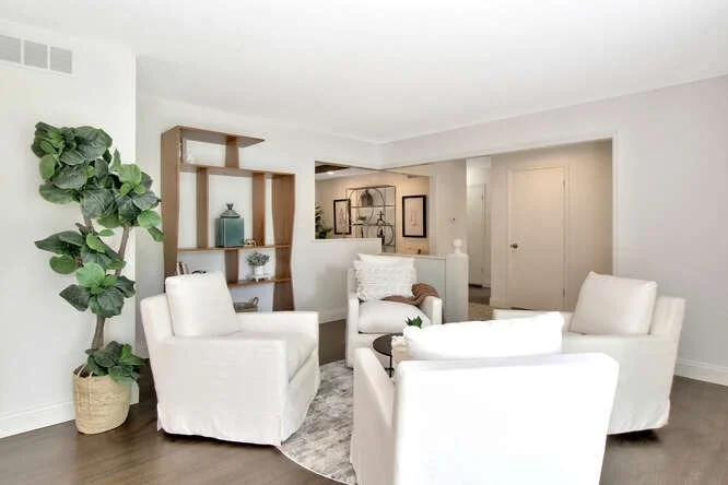 Living room with four white armchairs arranged around a round gray rug, a tall potted plant on the left, and built-in shelves with decorative vases on the left wall, neutral-colored walls, and hardwood floors.