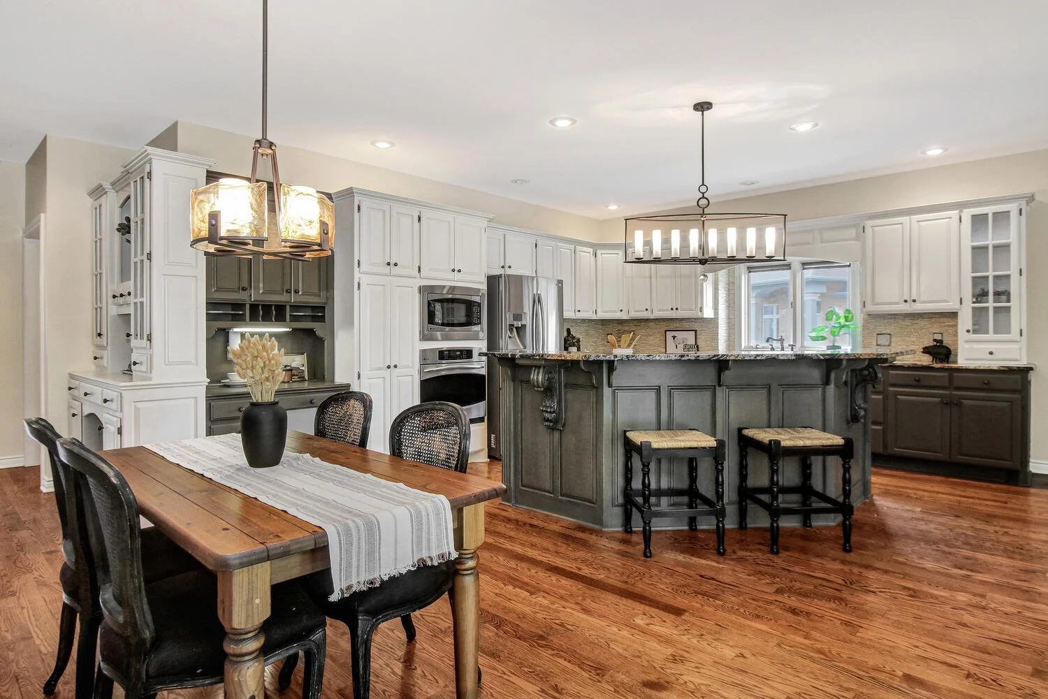 Open kitchen with white cabinets, a gray island with decorative corbels, and a wooden dining table with black chairs and a vase of flowers.