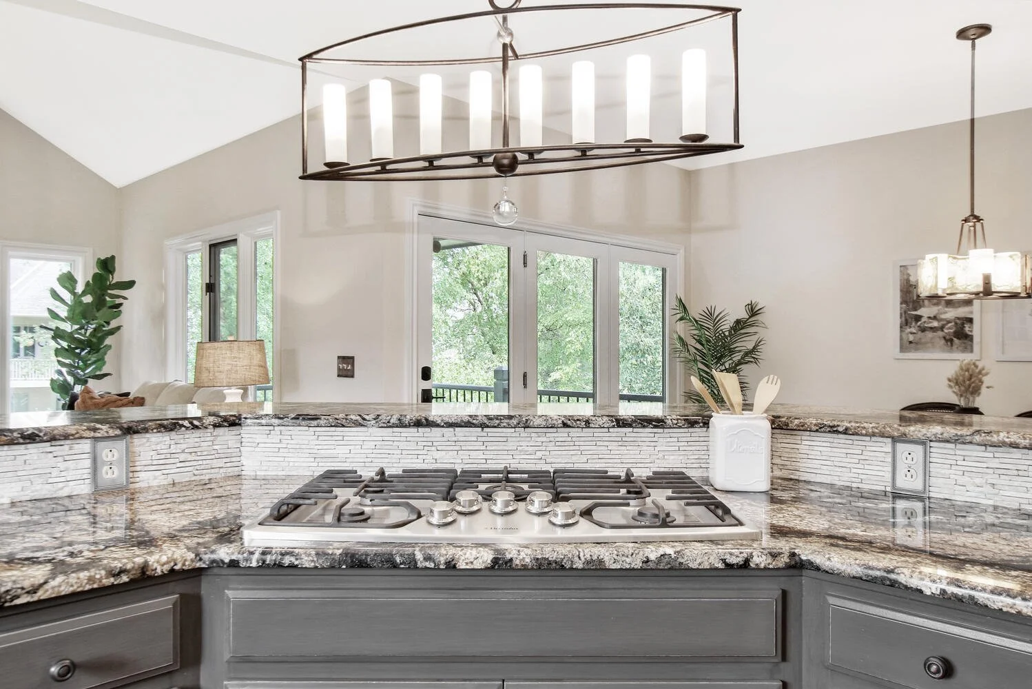 Kitchen with granite countertops, stovetop, gray cabinets, and a view of a living area with a lamp and plants through large windows.
