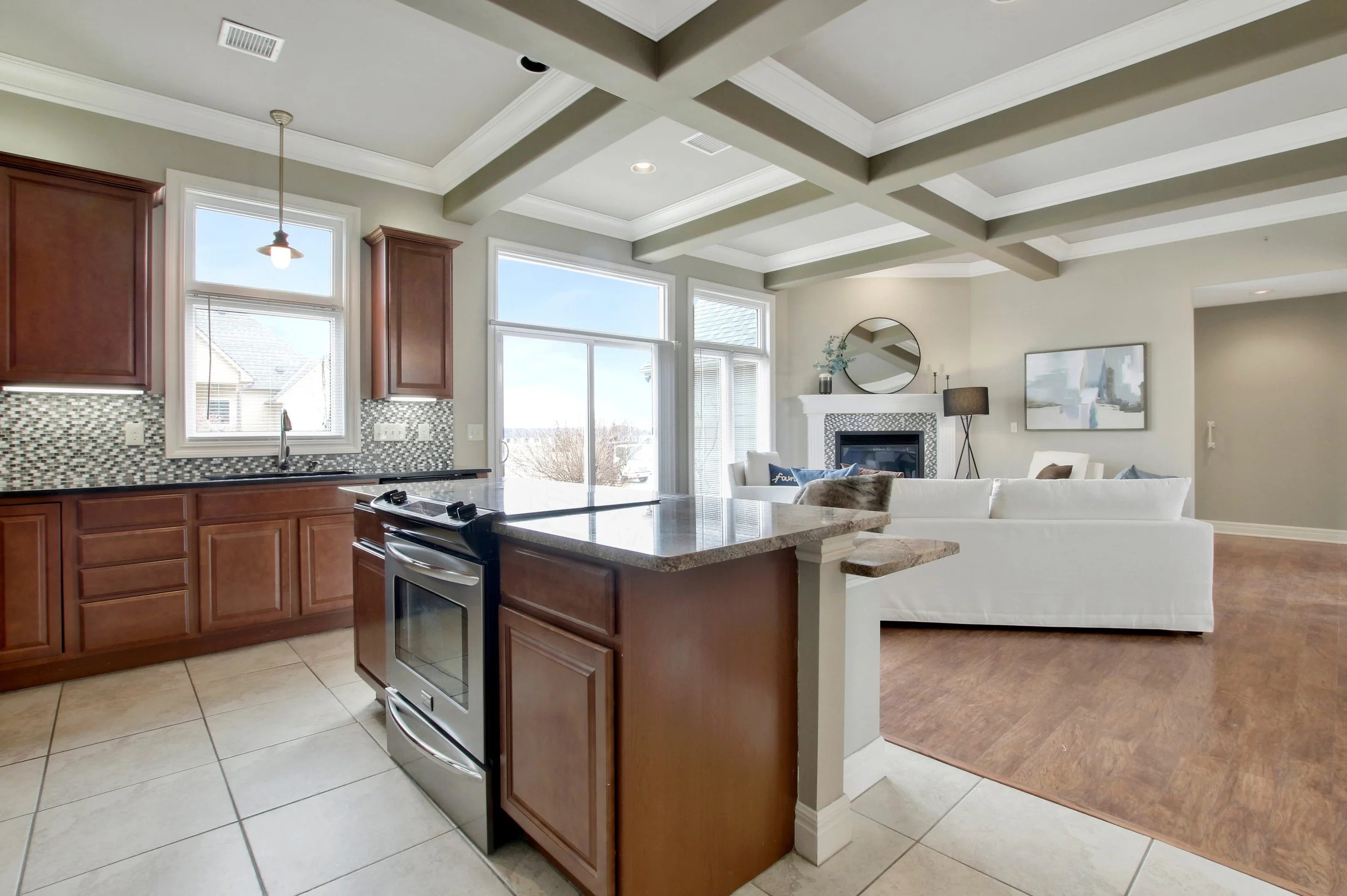 Open-concept kitchen and living room with large windows, brown kitchen cabinets, granite countertops, and a white sofa. The living room includes a fireplace with a round mirror above it, a floor lamp, and artwork on the wall.