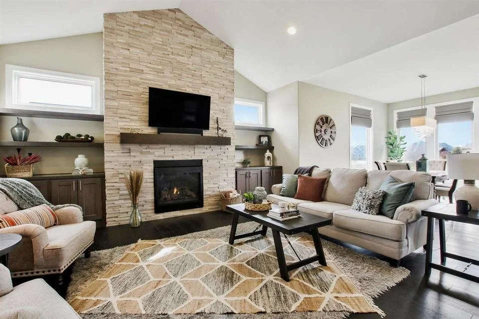 Modern living room with a stone fireplace, wall-mounted TV, beige sofa, armchair, wooden coffee table, and large windows