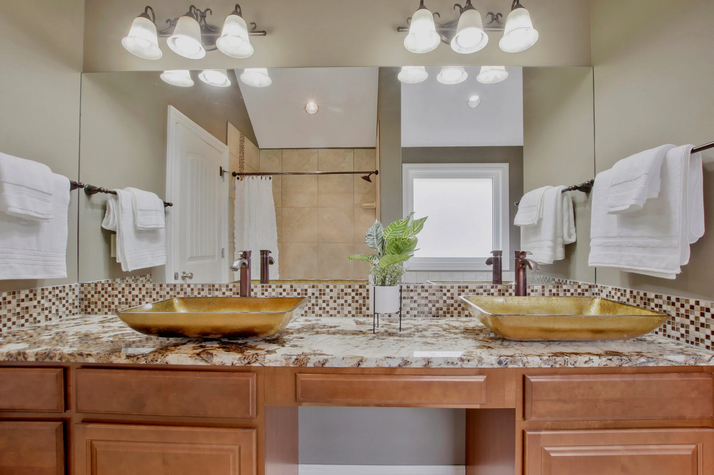 Bathroom with dual gold sinks, granite countertop, large mirror, wooden cabinets, white towels, potted plant, and a window with a frosted glass panel.