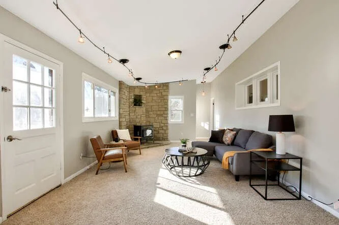 Living room with a gray sofa, black side table and lamp, armchairs, a stone fireplace, and windows letting in natural light.